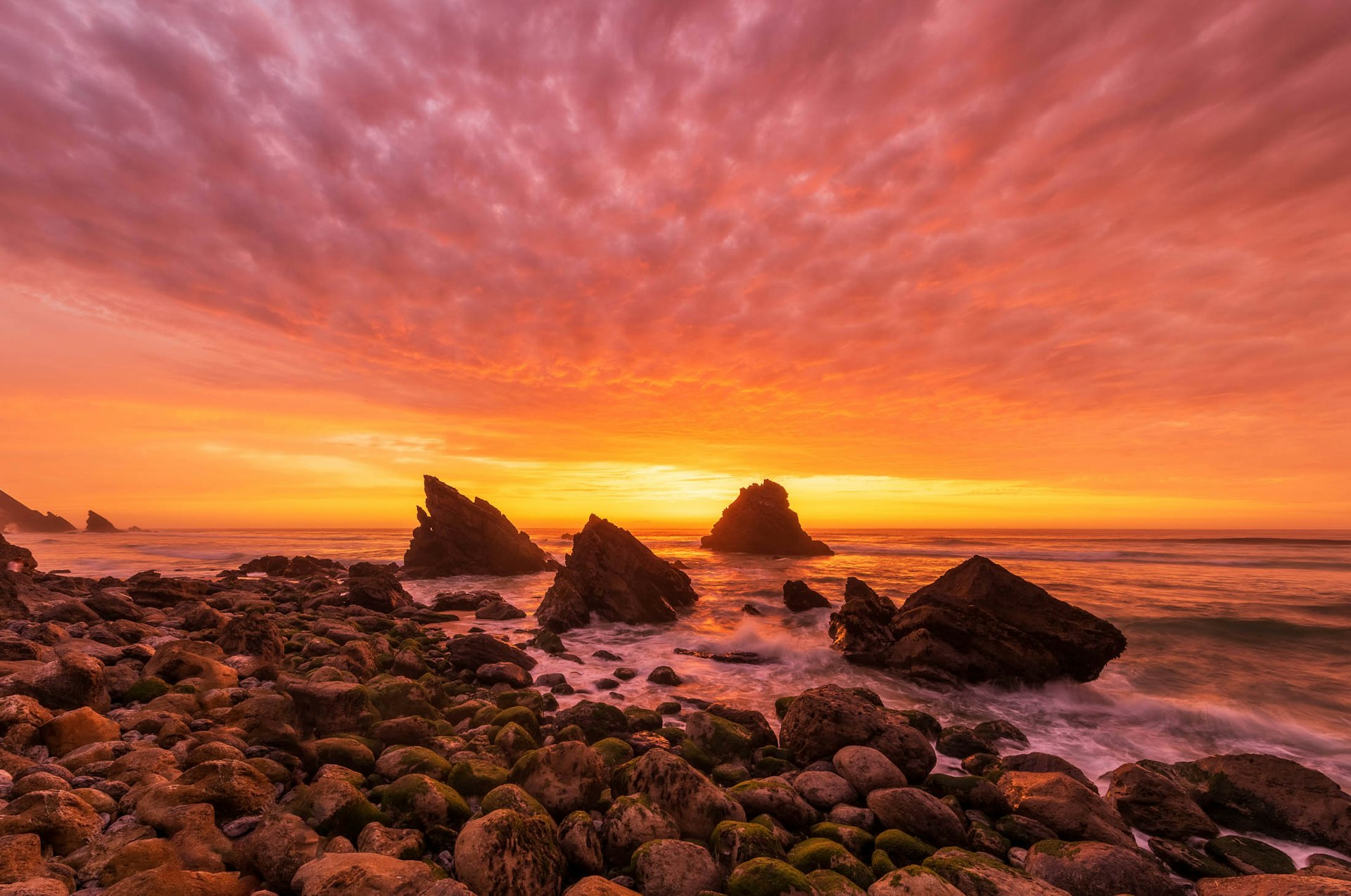 A dramatic sunset streaks orange and pink light across the sky with rocks on a beach in silhouette 
