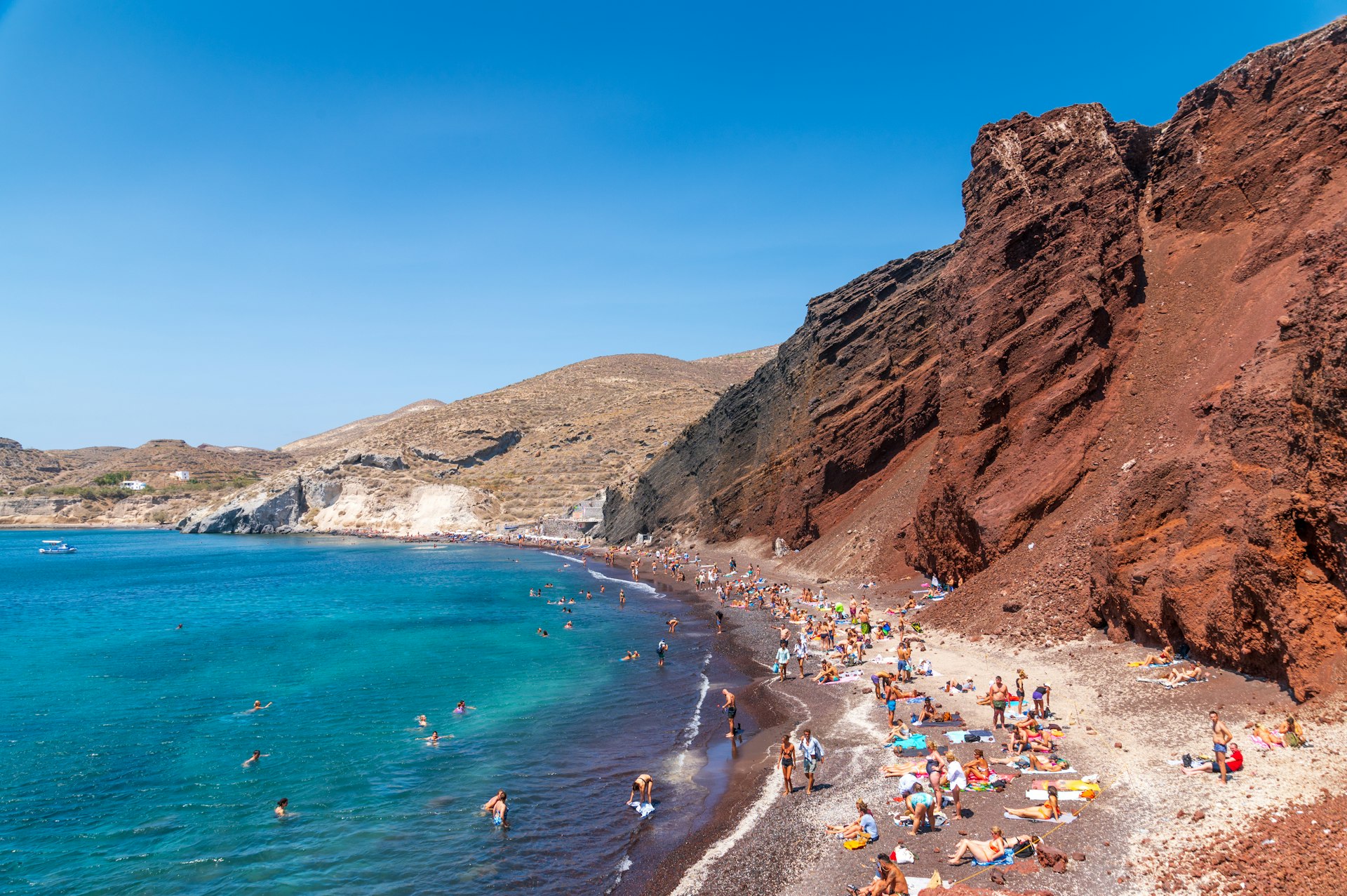 People line the narrow sands of a beach below striking red cliffs