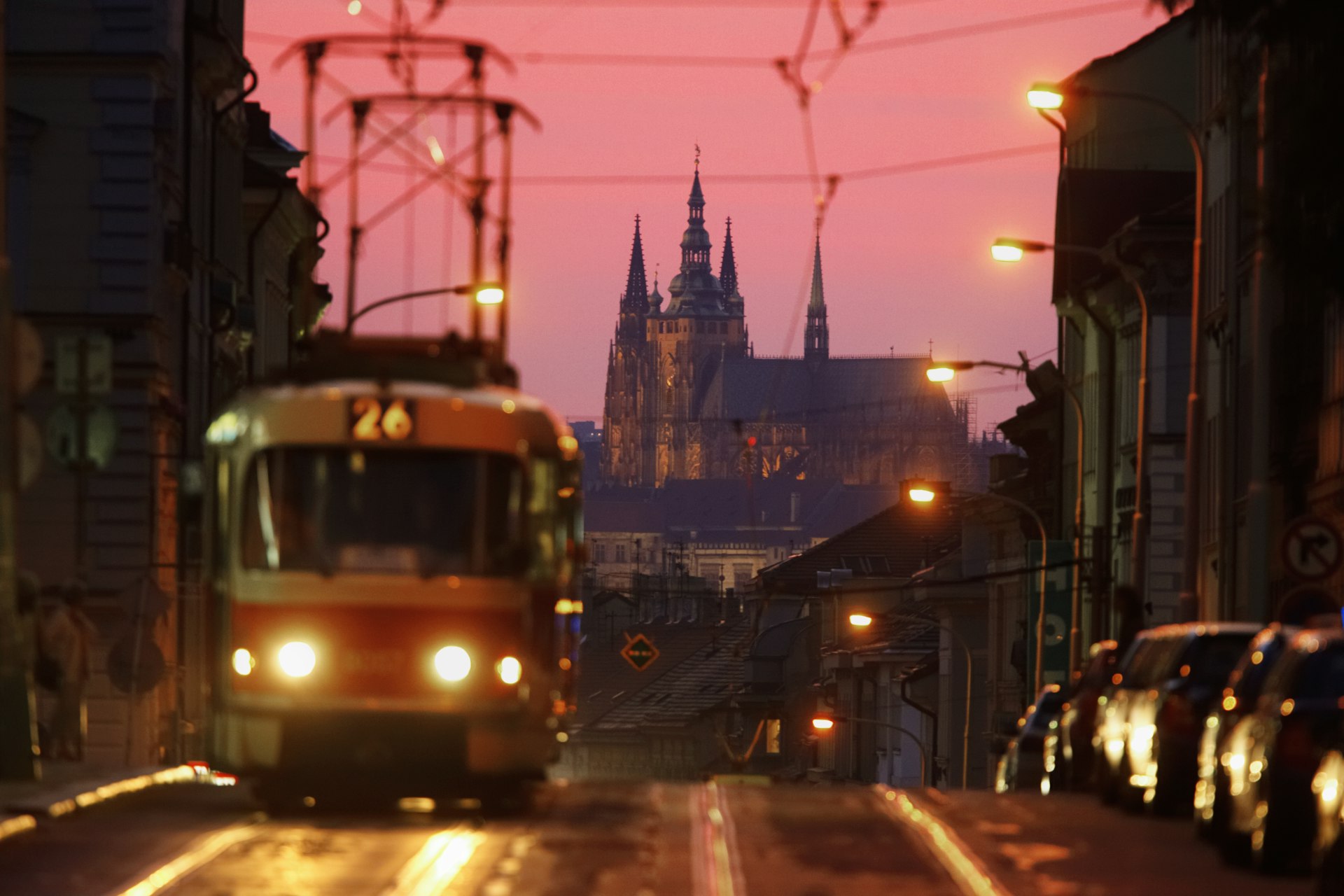 A tram runs along a street at dusk. A huge multi-spired church building is silhouetted in the distance