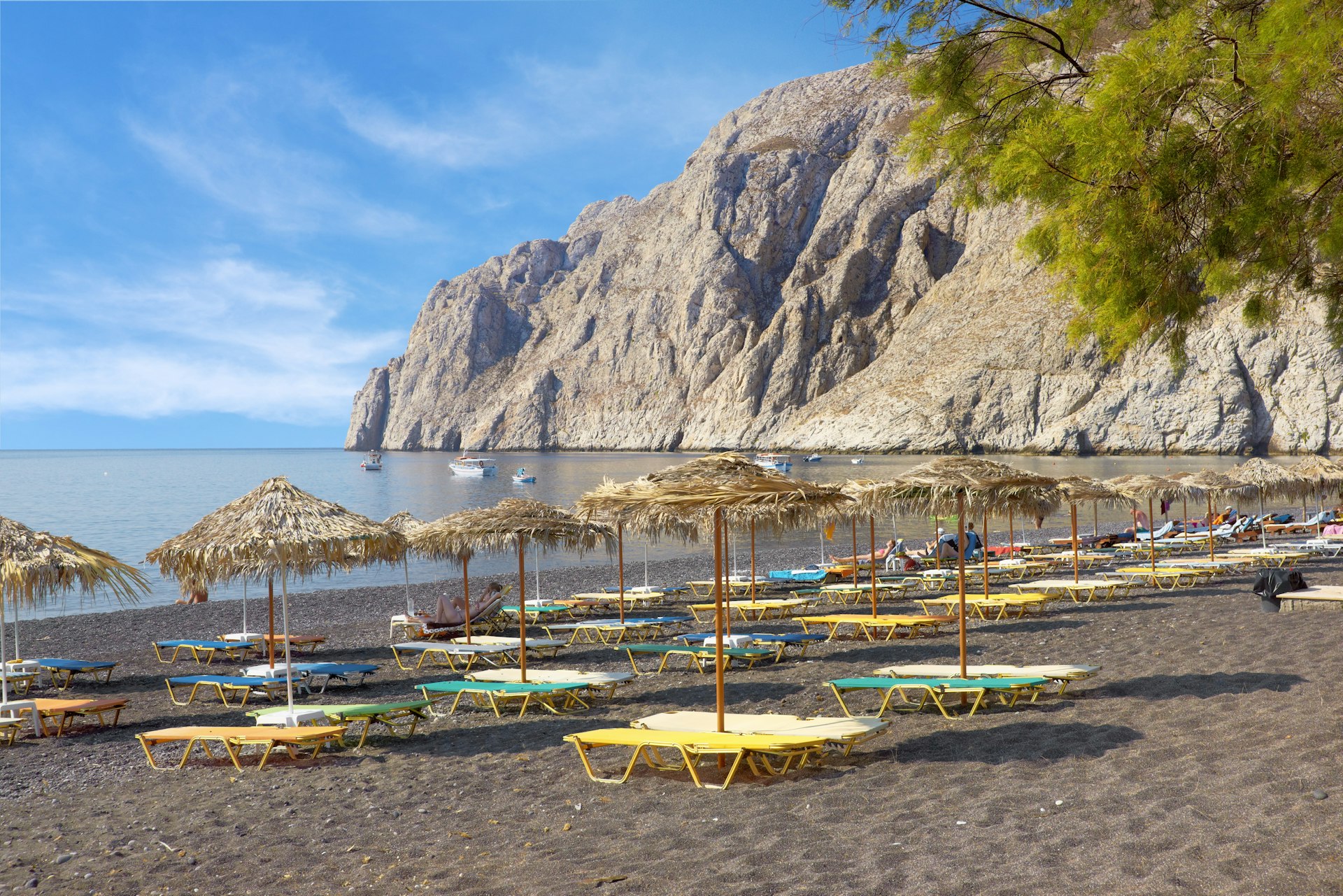 A series of sun loungers under palm-tree umbrellas on a beach with dark sand