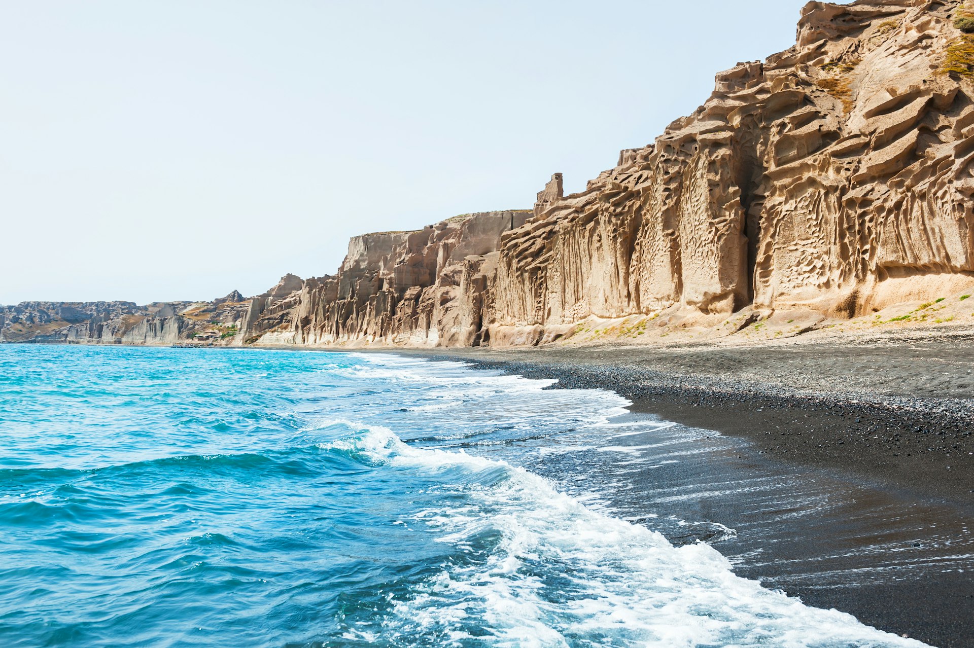 The sea laps on the shores of a black-sand beach, with sandstone cliffs towering over it