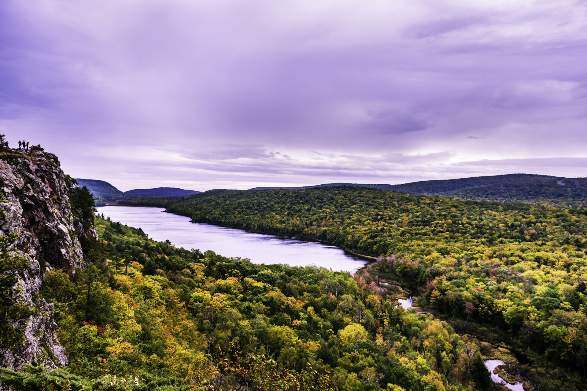 A viewpoint over a lake surrounded by woodland. A small group of hikers stand to the left on a rocky outcrop