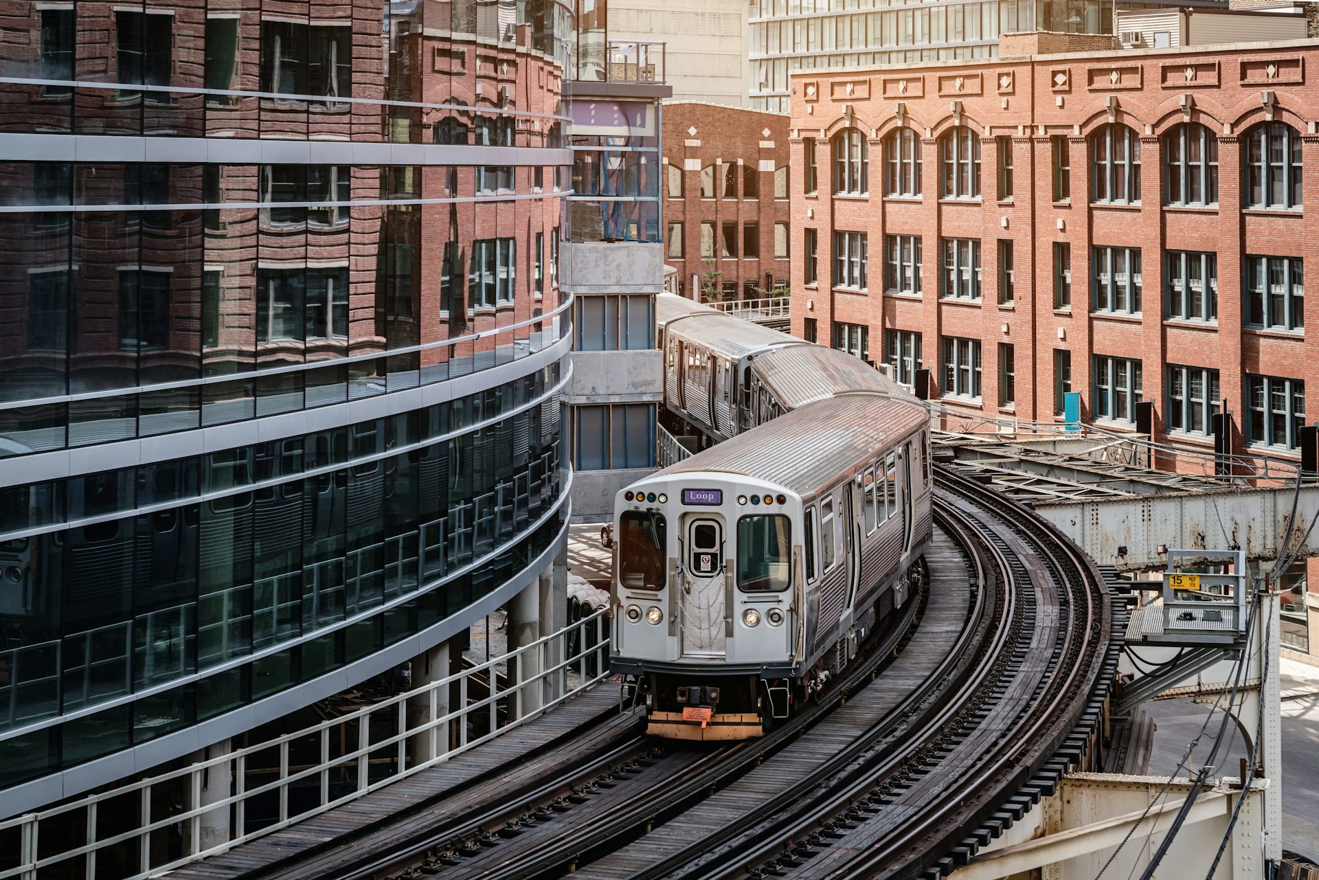A silver commuter train runs on an elevated track between buildings