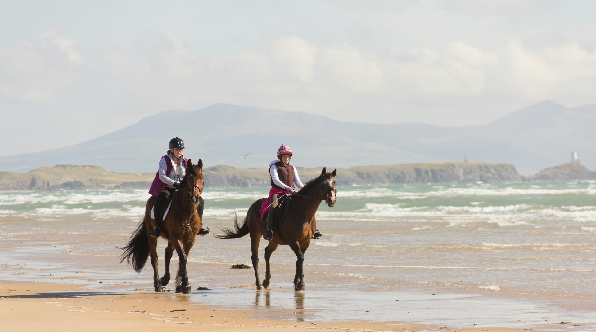 Two people ride on brown horses on an otherwise empty sandy beach