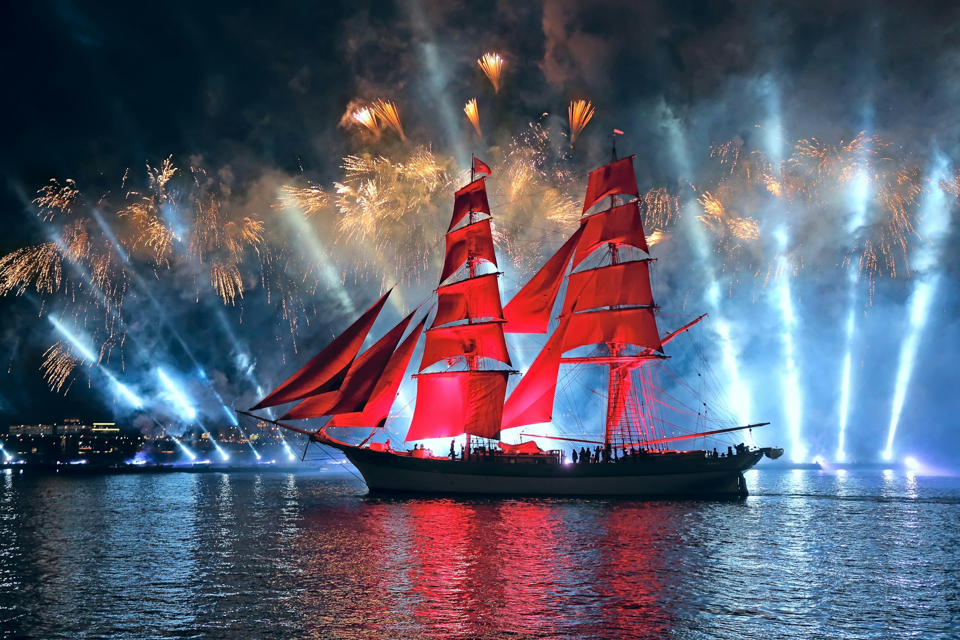 A sail boat on a river at night. It has large red sails and is backlit by bright lights