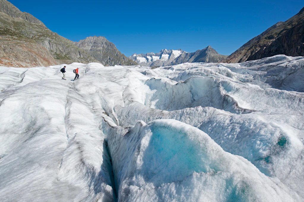 Two hikers walk over the Aletsch Glacier, a rugged icy landscape. Above them the sky is bright blue.