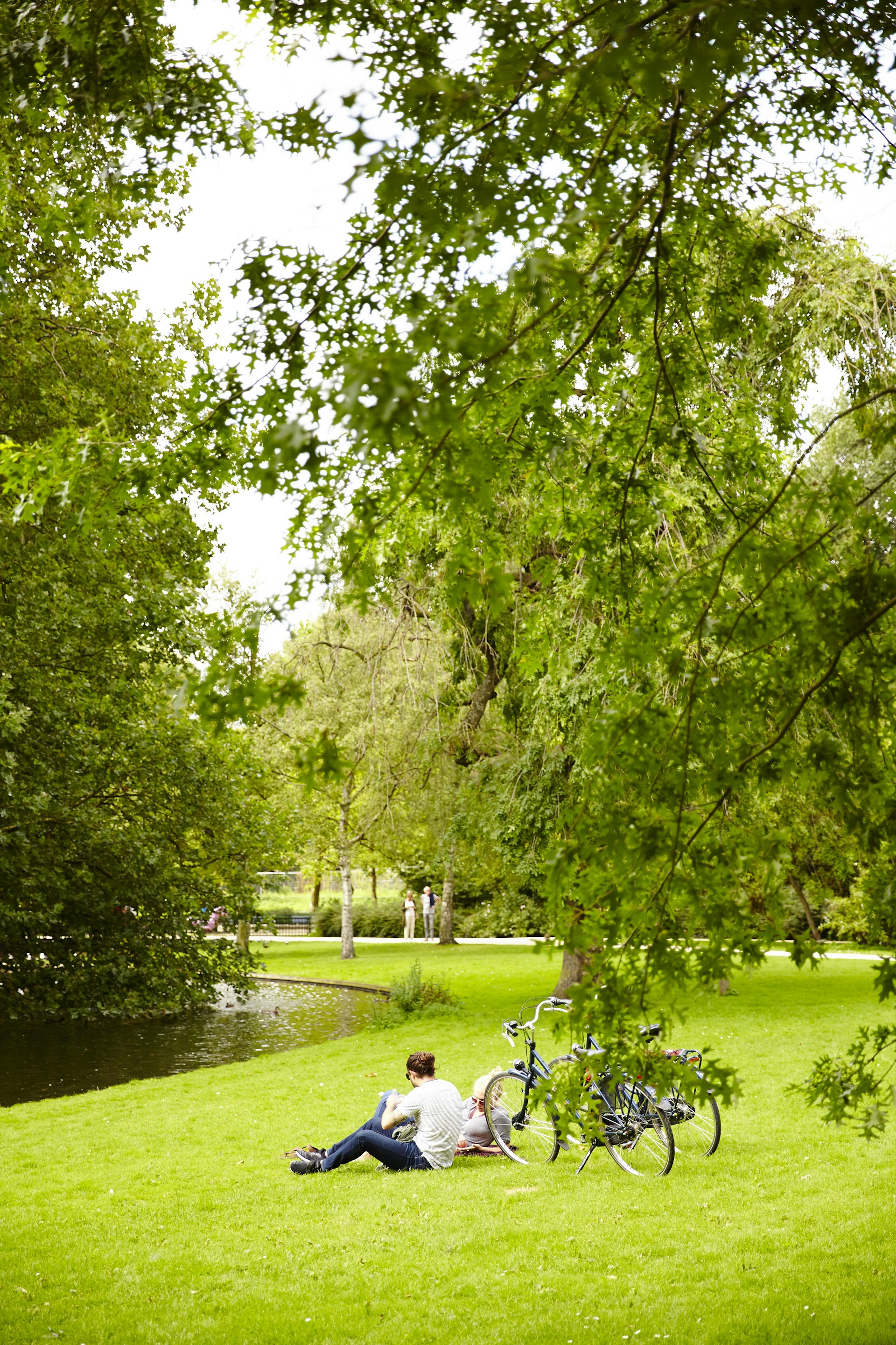 A couple lay on the grass near their bikes in parkland with a small stream running nearby