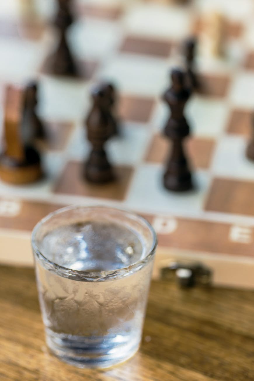 A small glass of a clear liquid with a chess board in the background