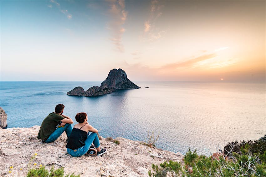 Two people sit on a rocky outcrop looking out to sea at an islet with the sun setting behind it