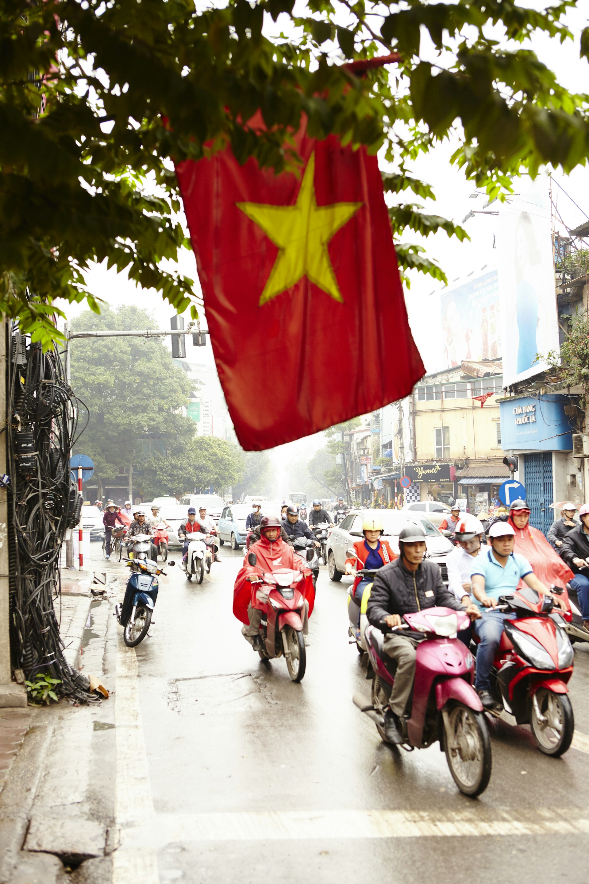 A red flag with a yellow star in the center hangs low over a street packed with motorcyclists
