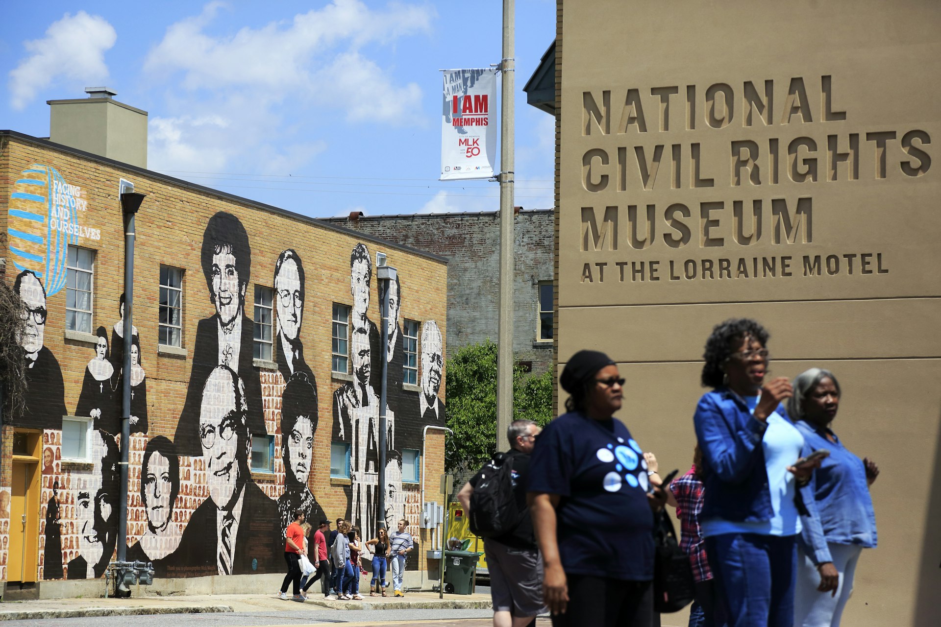 A trio of women walk towards the National Civil Rights Museum at the Lorraine Motel. In the background you can see a brick building covered in paintings of prominent Americans. 