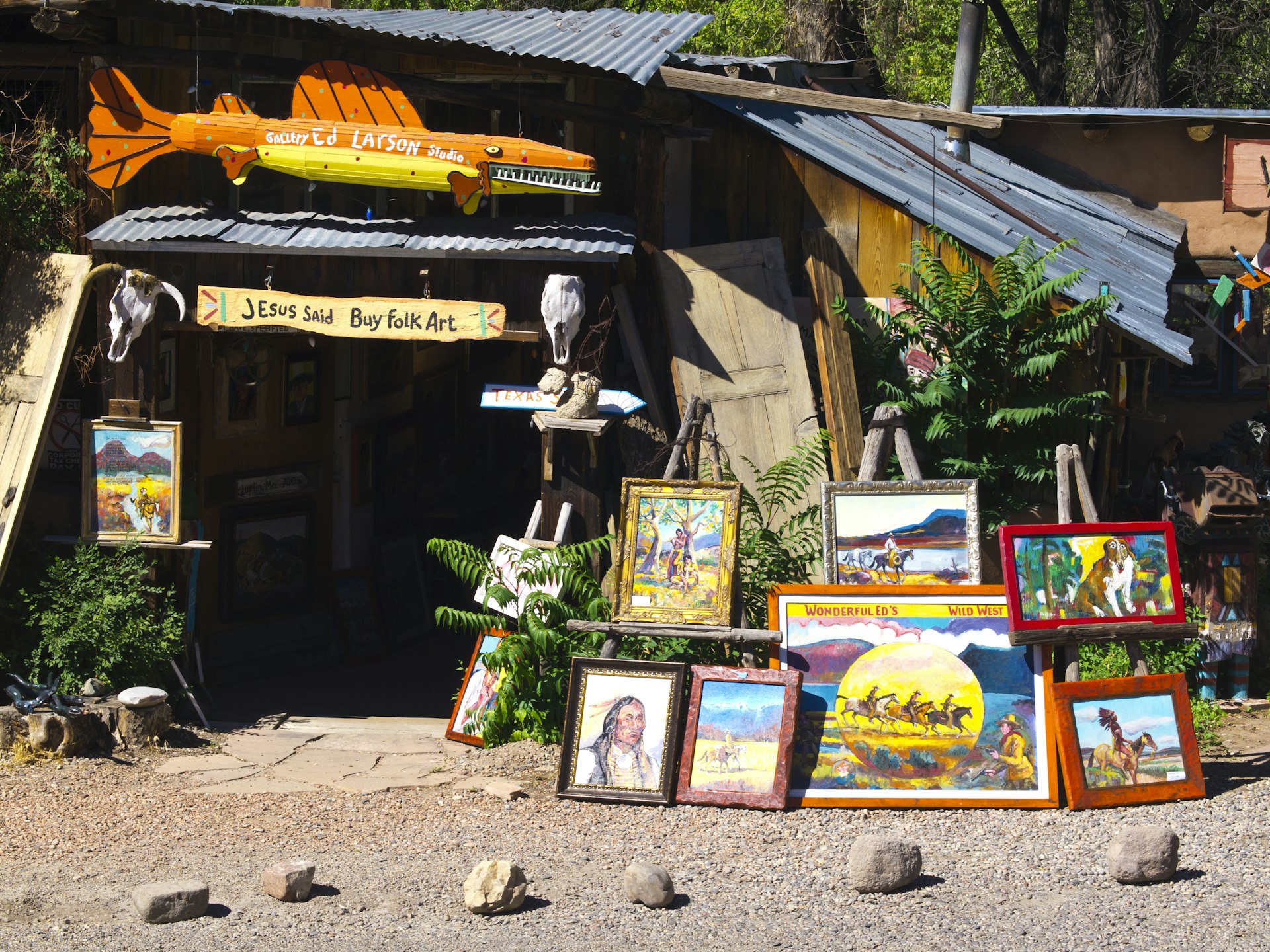 Ed Larson studio and gallery in a very old adobe and wood farm house with commercial sign inviting people to by folk art. In front of the gallery on the sun there are some paintings with motives from Wild West.