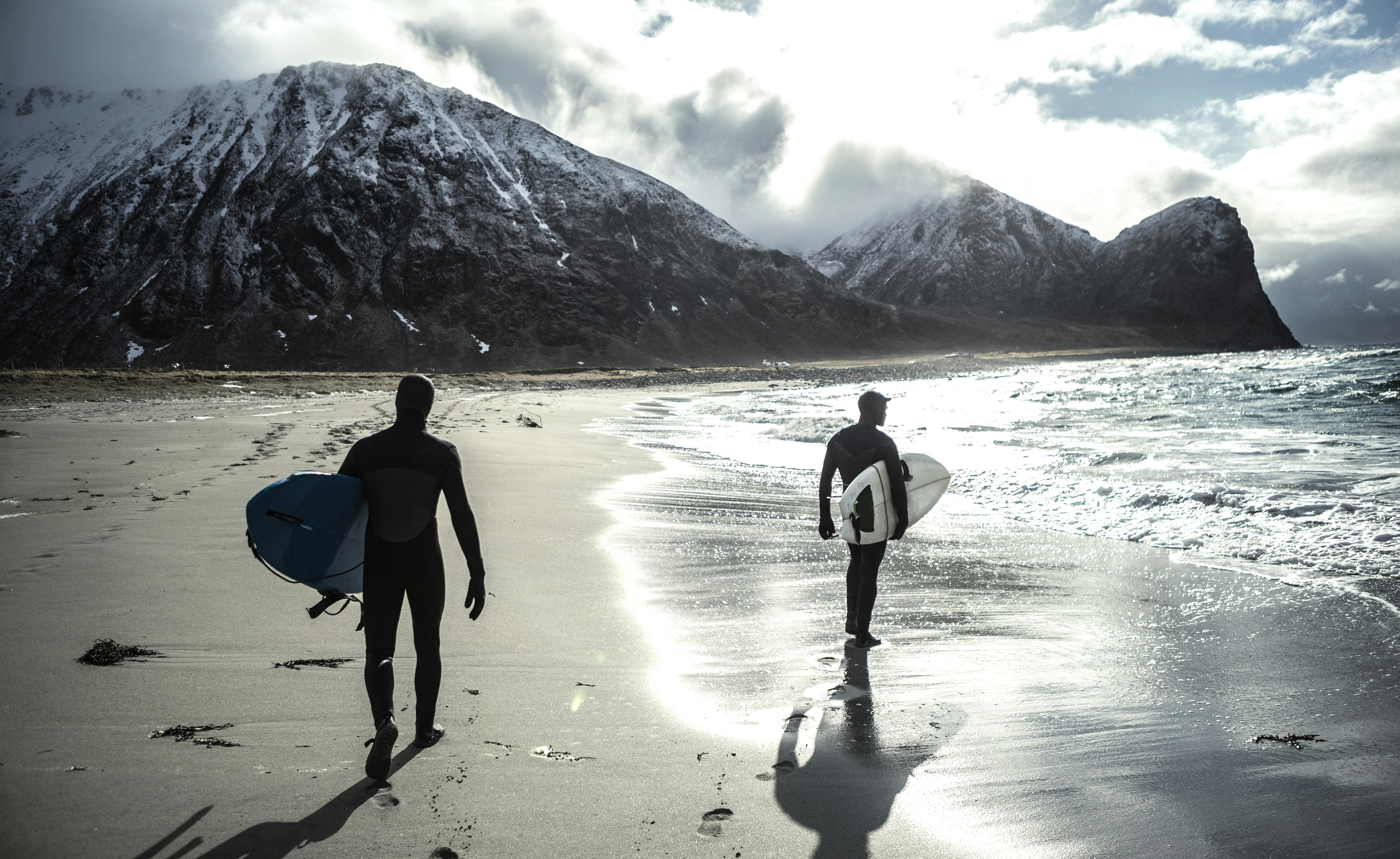 Two surfers wearing wetsuits and carrying surfboards walk along a beach in Norway with snow-capped mountains behind.