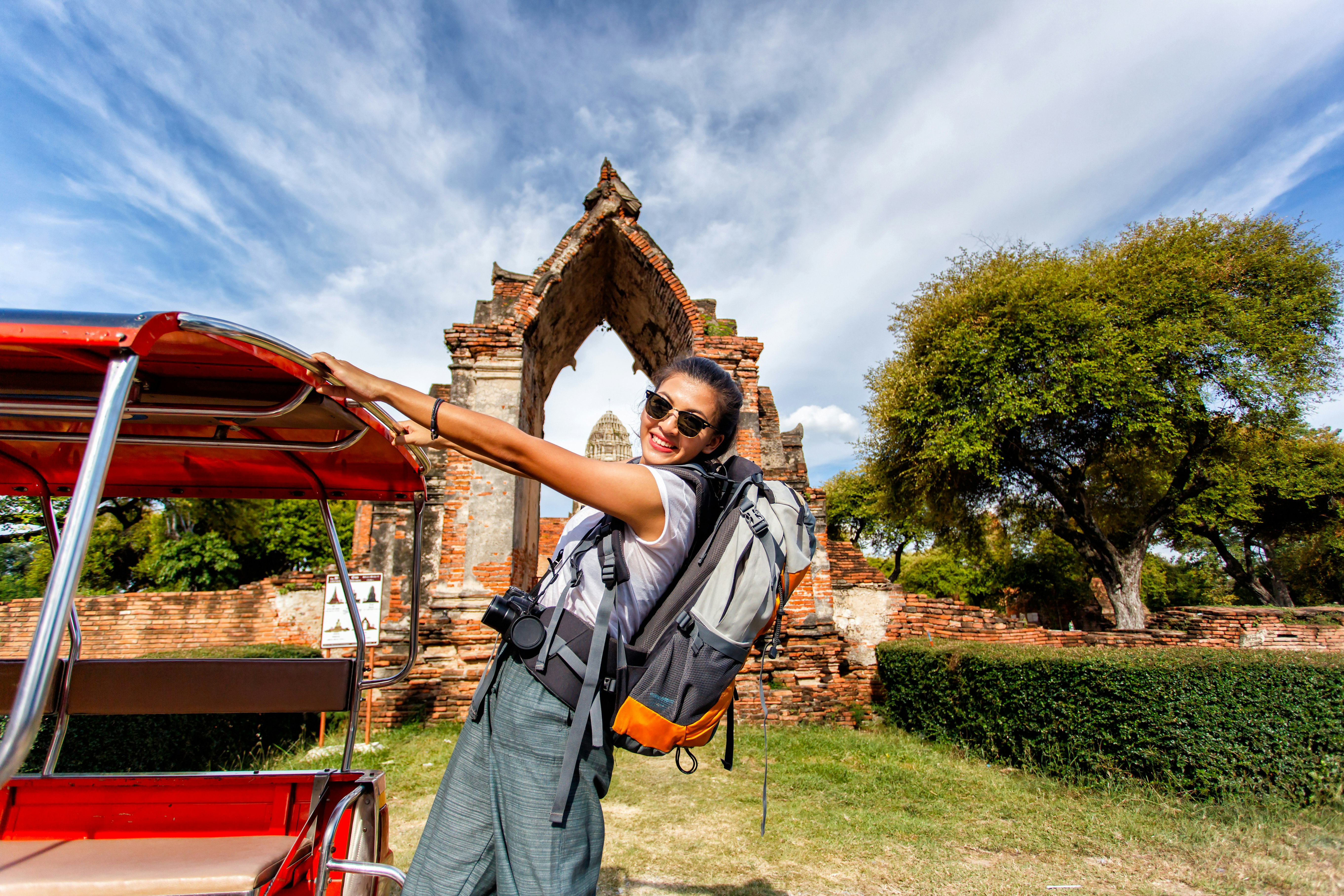Young asian female traveler with backpack standing on Tuk Tuk with Wat Mahathat in background