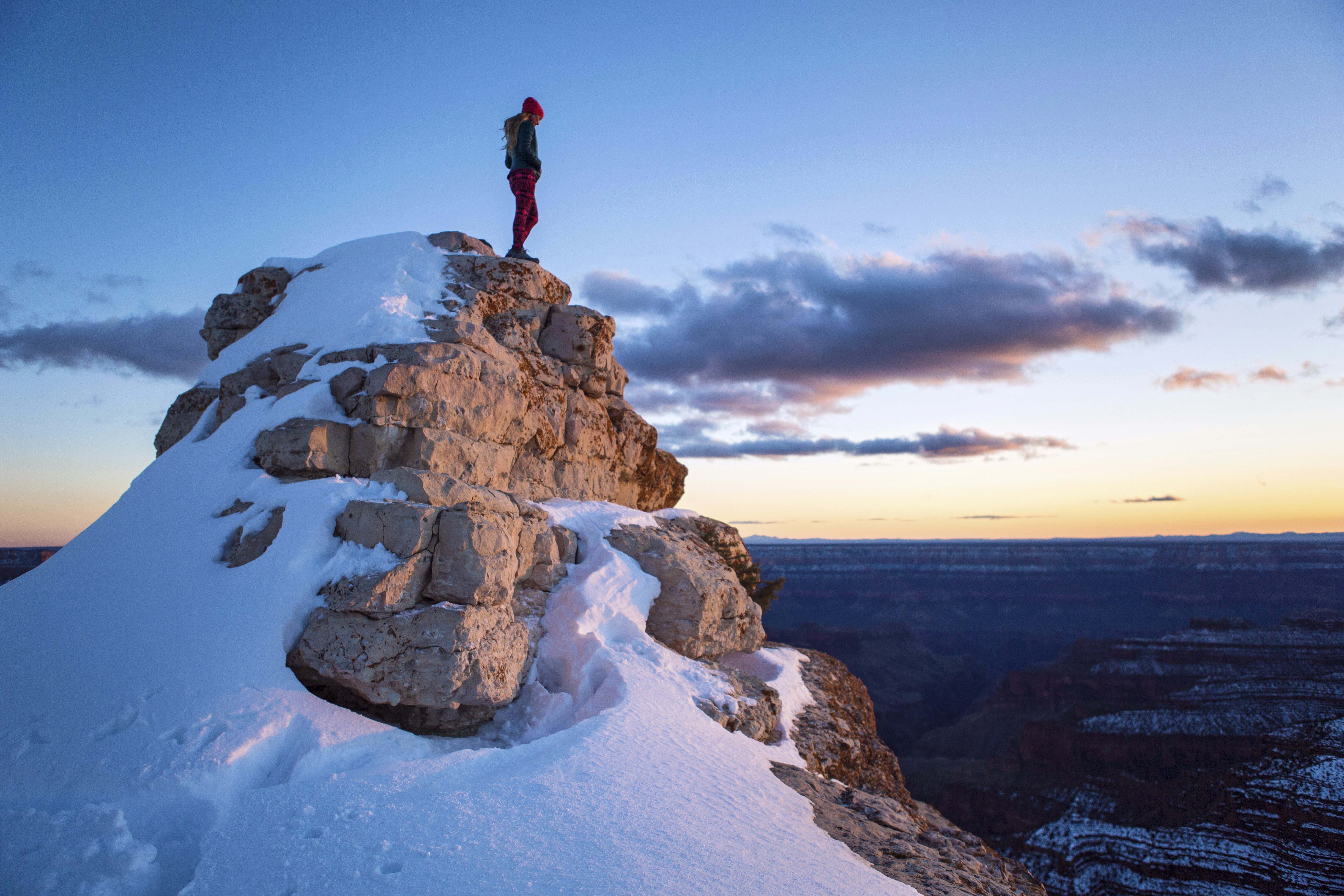 A fit, female hiker stands atop a snow-covered rocky high point while bathed in the colors of sunset at the North Rim of the Grand Canyon. 