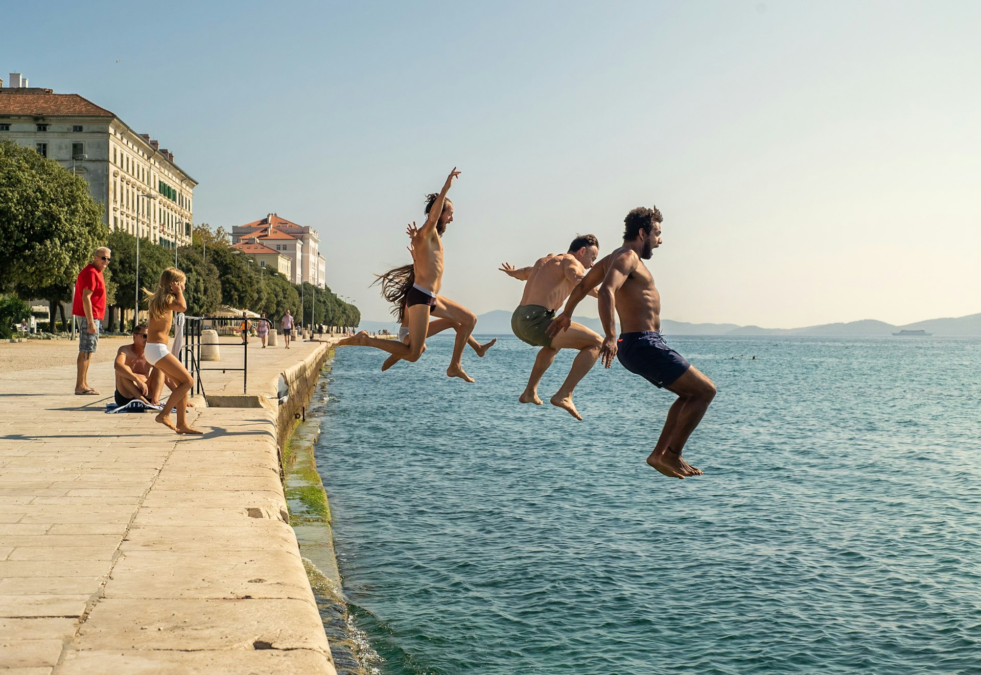 A group of people run off a walkway beside the sea and jump into the ocean