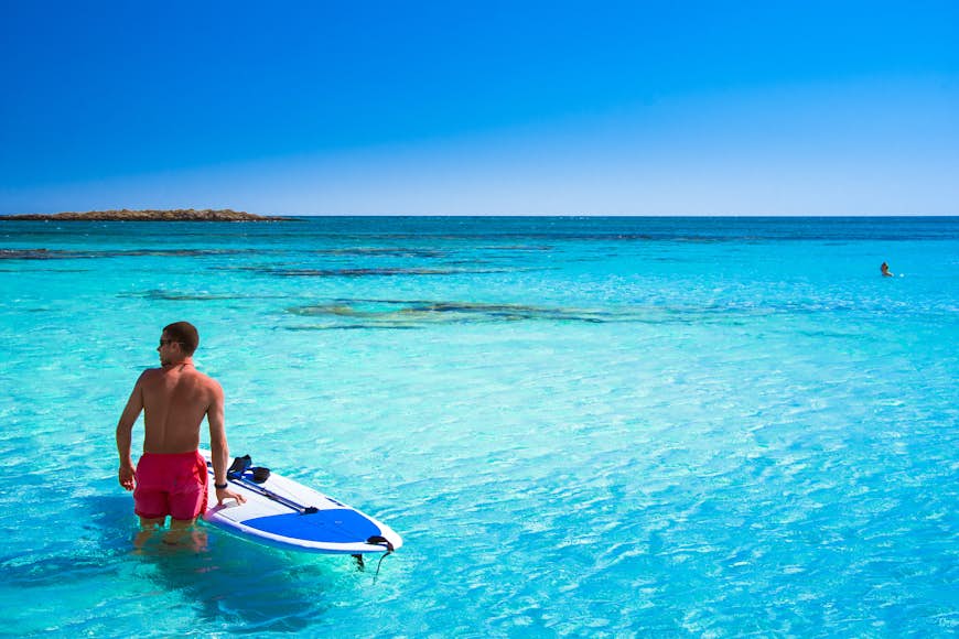A man with a surfboard standing in the turquoise water in Crete, Greece