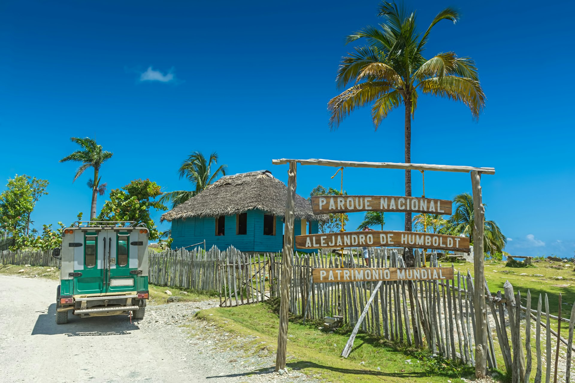 A sign at the entrance to Sign at Alejandro de Humboldt National Park in Cuba, with picket fence, thatched-roof visitor center, palm trees and blue skies