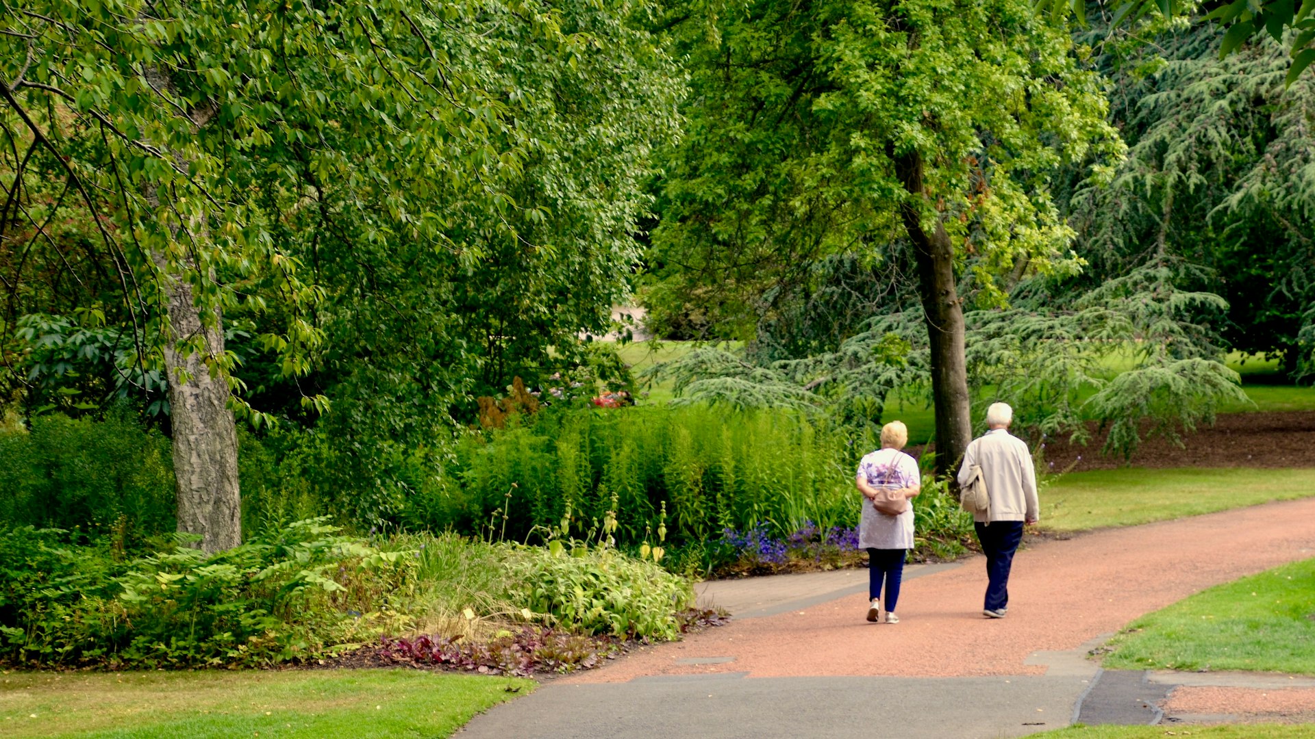 An elderly couple with their backs to the camera walk down a path through pristine gardens