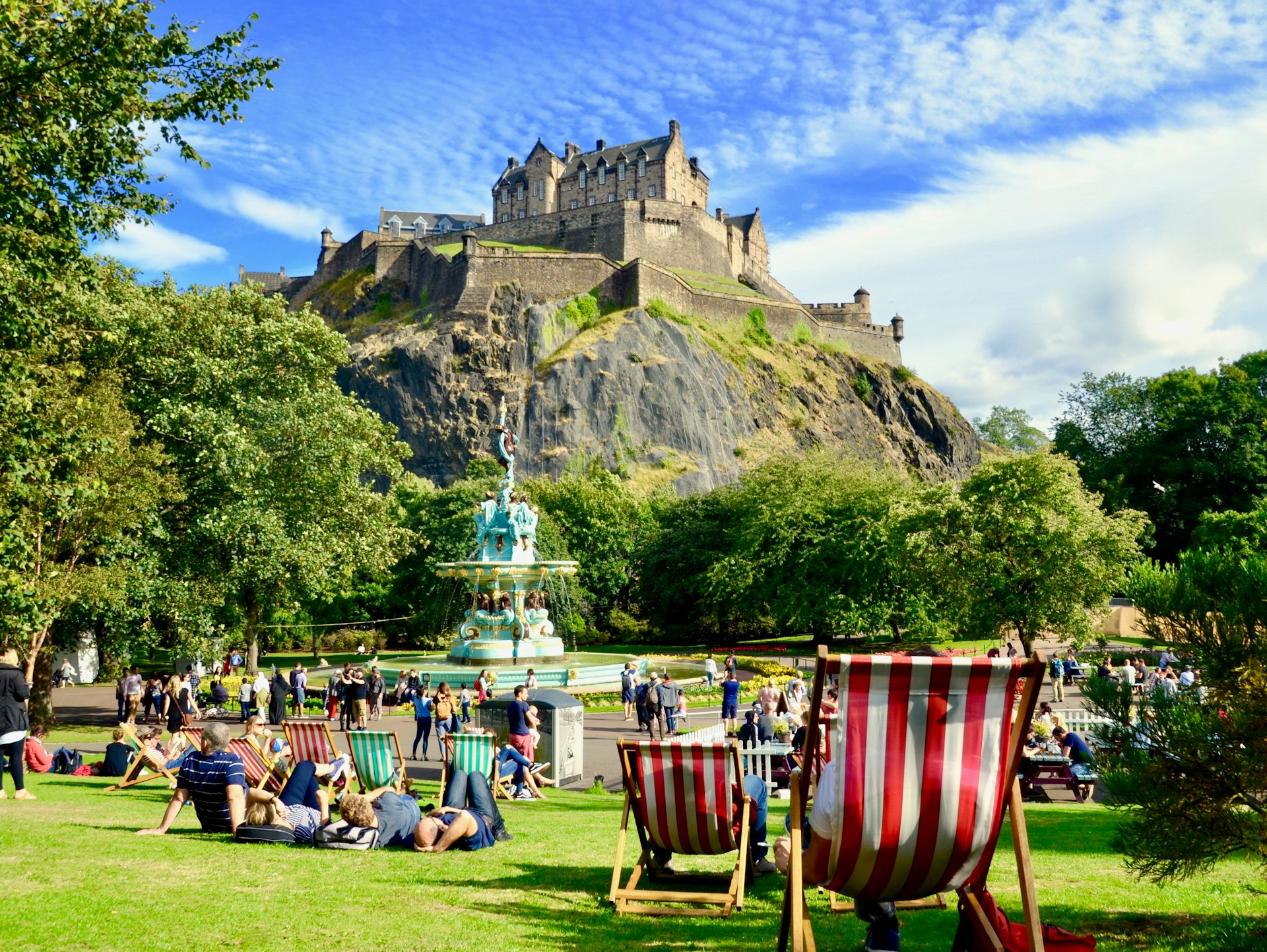 People sitting on the grass and relaxing in striped lawn chairs near Ross Fountain in Princes Street Gardens, with Edinburgh Castle above.
