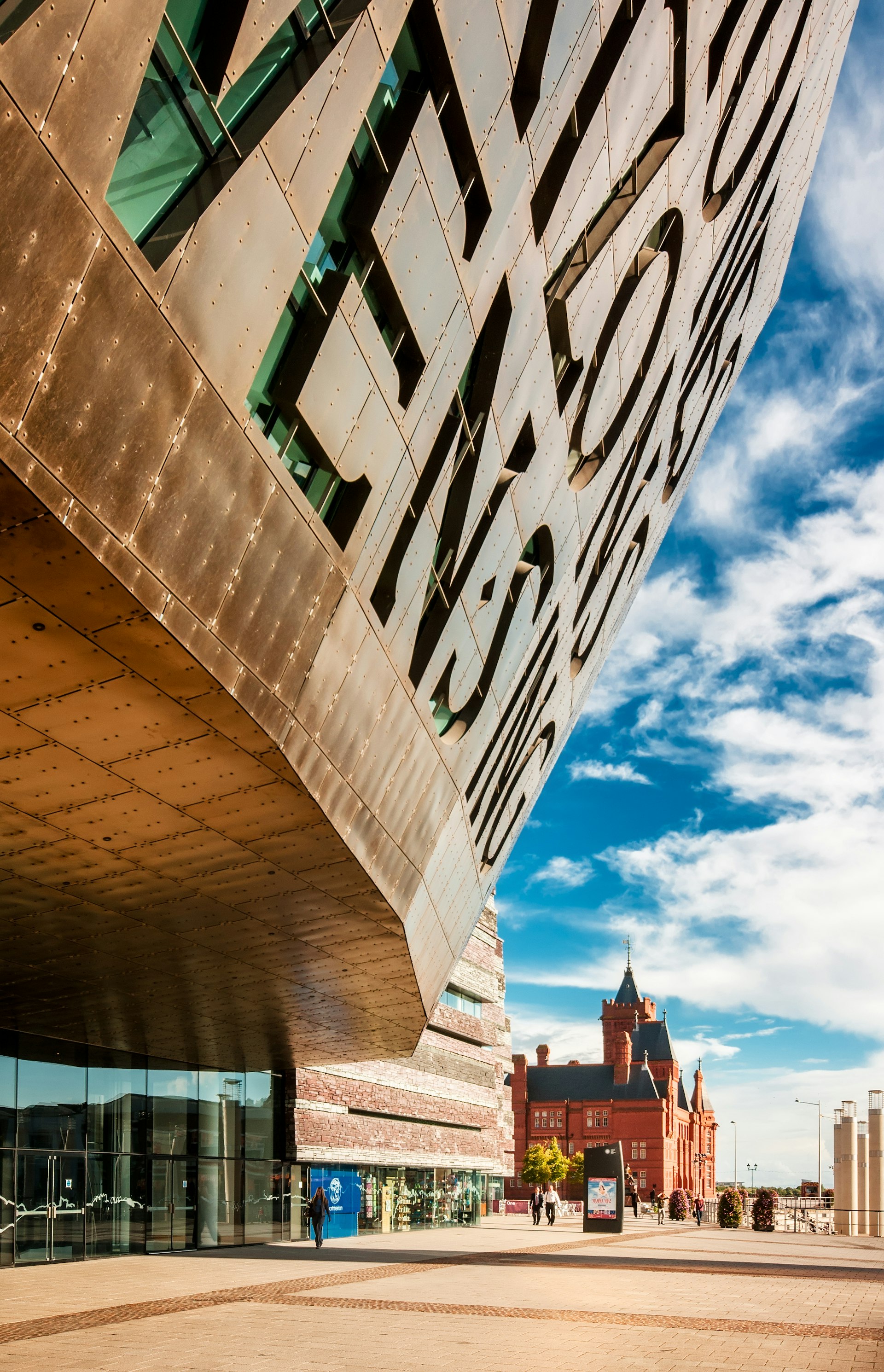 A large curved building with a copper facade. A red-brick church-like building stands in the background 