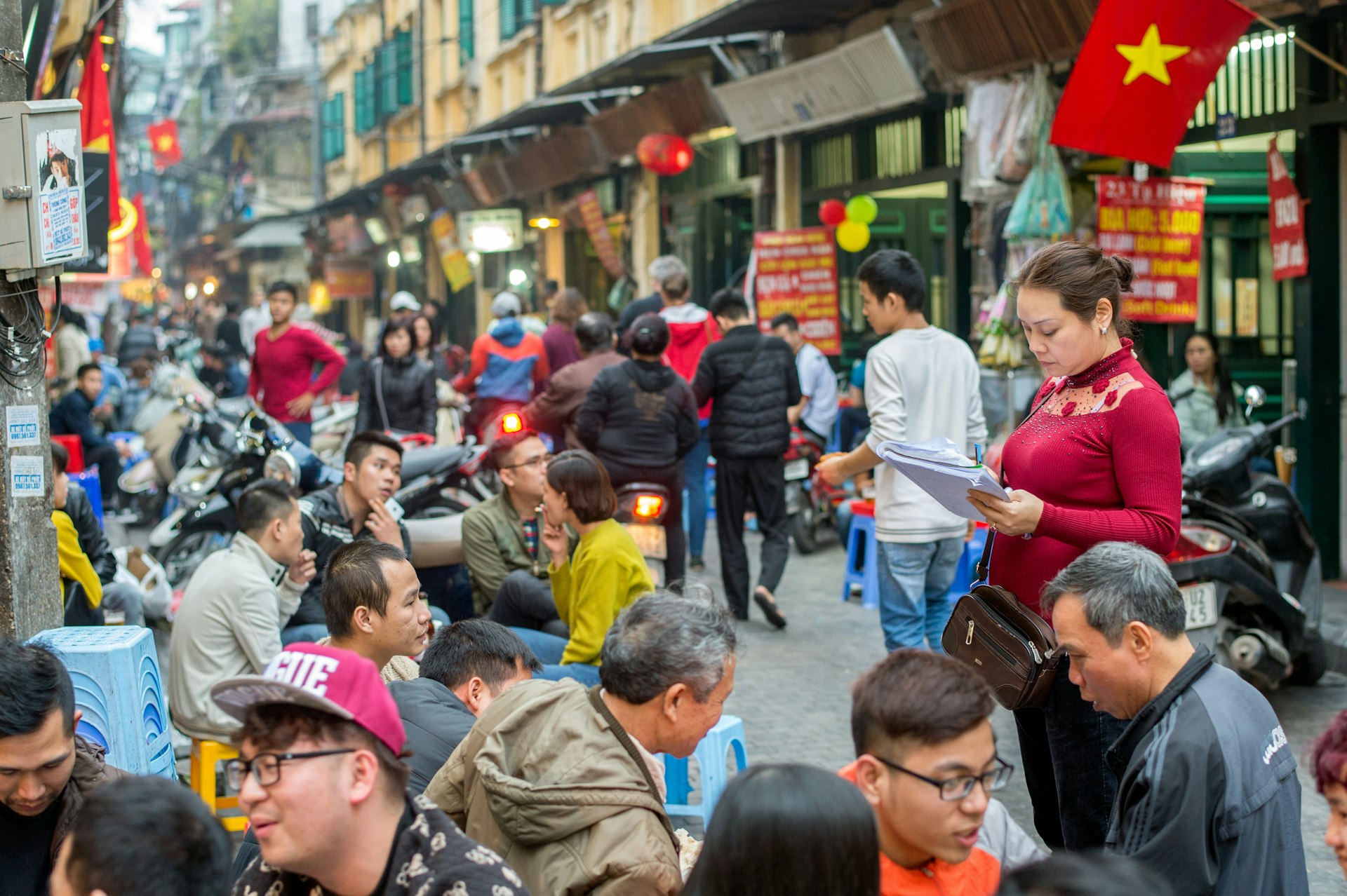 A street scene with many customers sat at stools outside a restaurant on the edge of the road. Vietnamese flags (red with a yellow star) hang along the street.