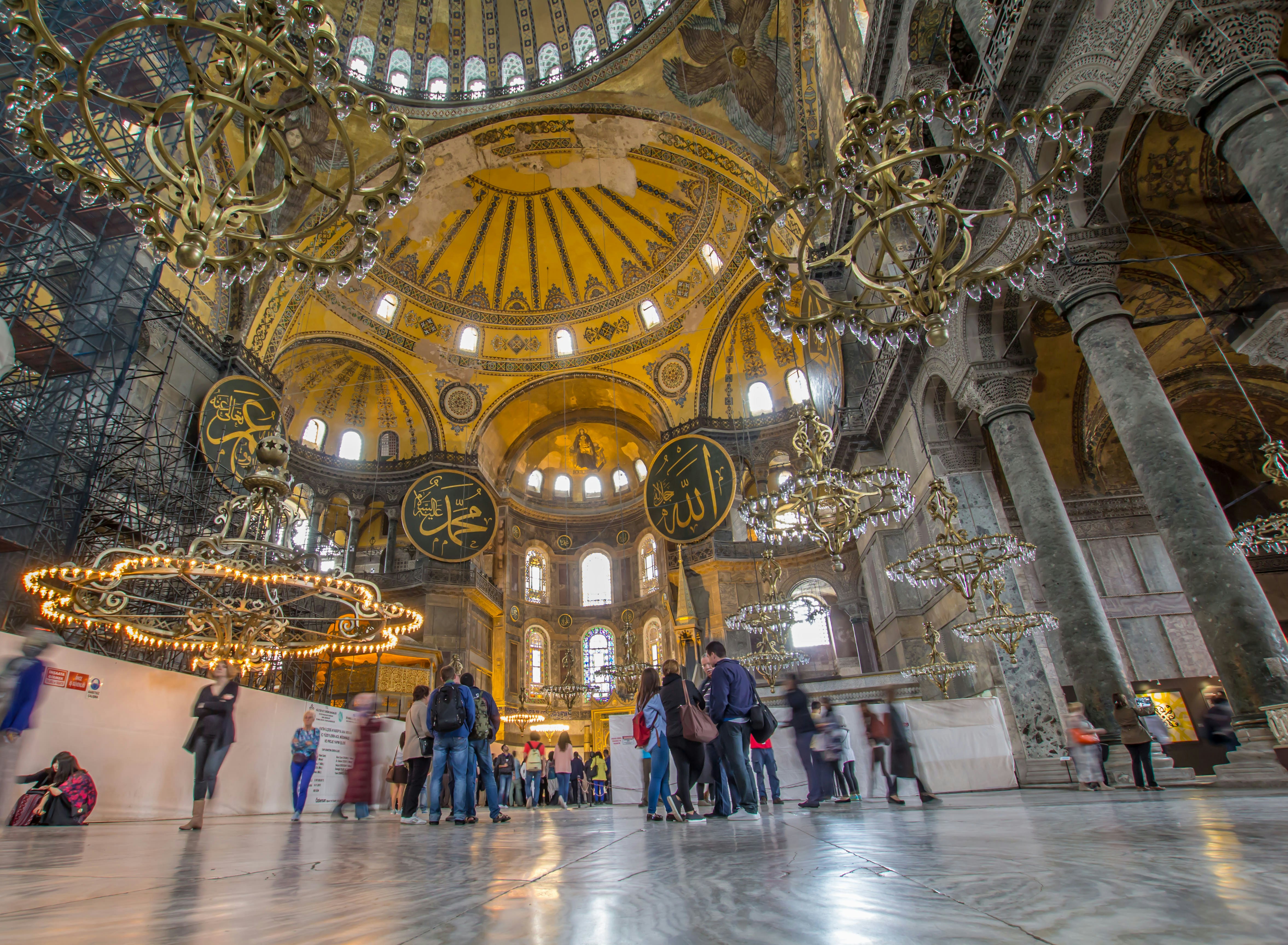 The interior of the Hagia Sophia in Istanbul, with a huge central golden dome and massive chandeliers hanging down from the ceiling.