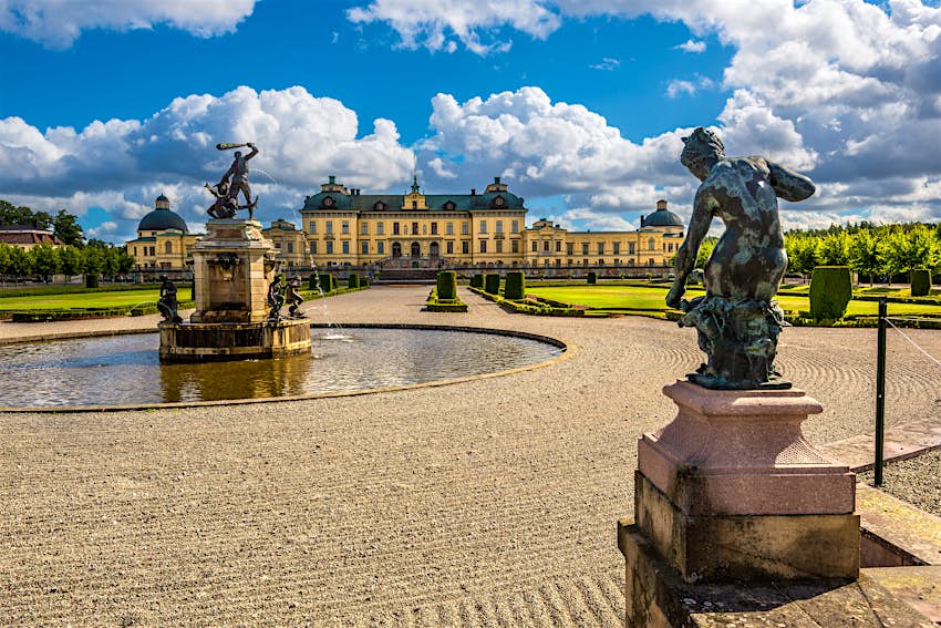 Fountains and statues in manicured grounds with a vast pastel-yellow-colored palace in the distance