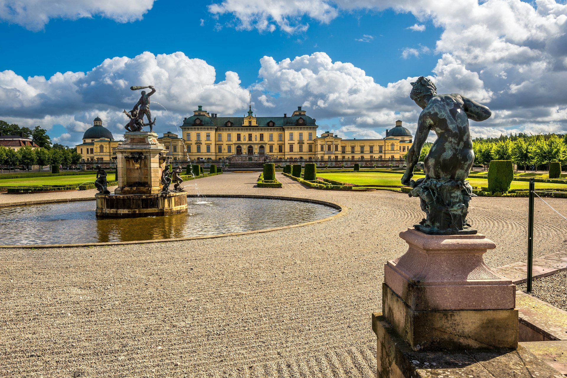 Fountains and statues in manicured grounds with a vast pastel-yellow-colored palace in the distance
