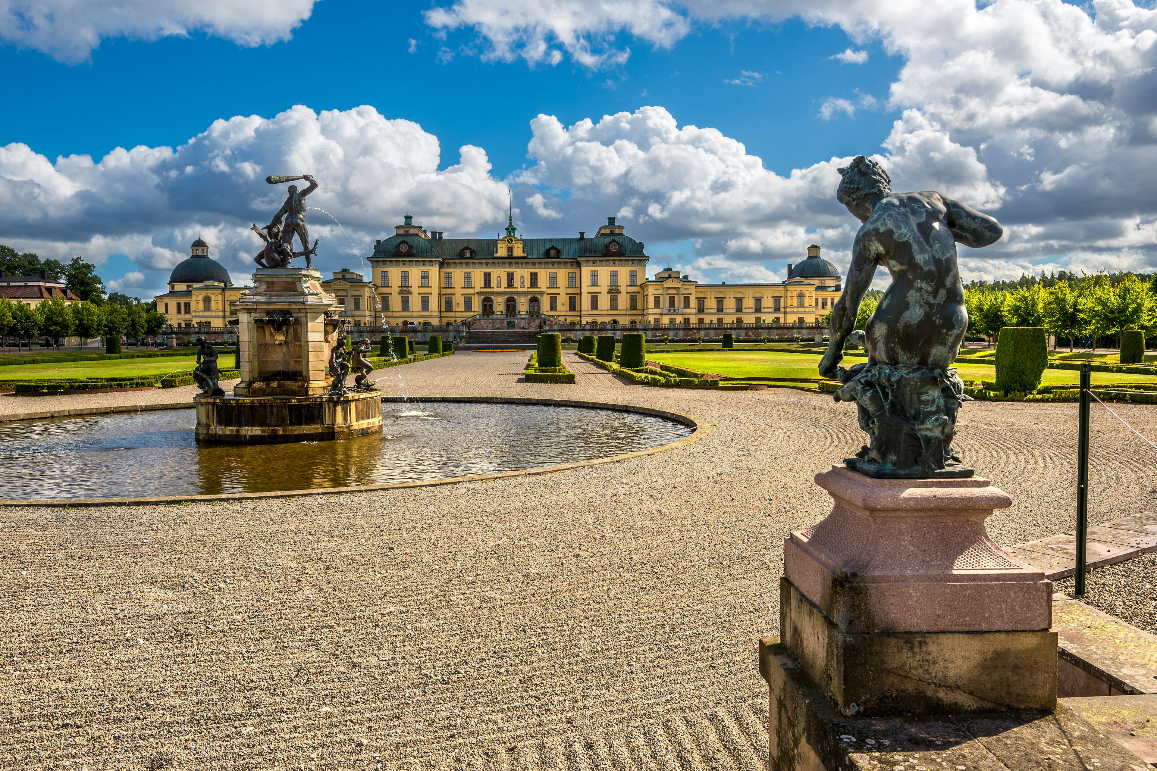 Fountains and statues in manicured grounds with a vast pastel-yellow-colored palace in the distance