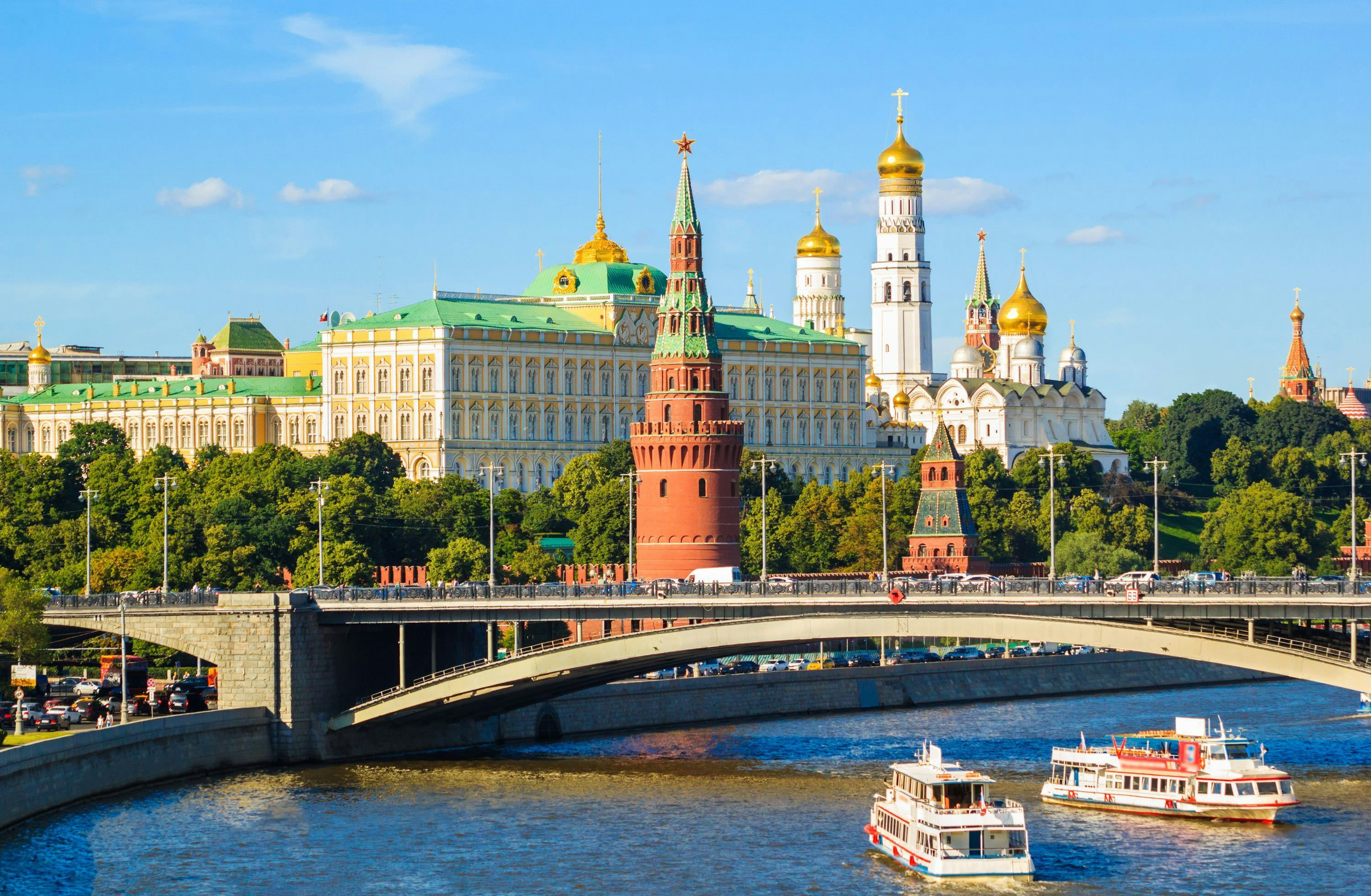 A series of large buildings, the outer one with dark red walls, line a riverside in a city