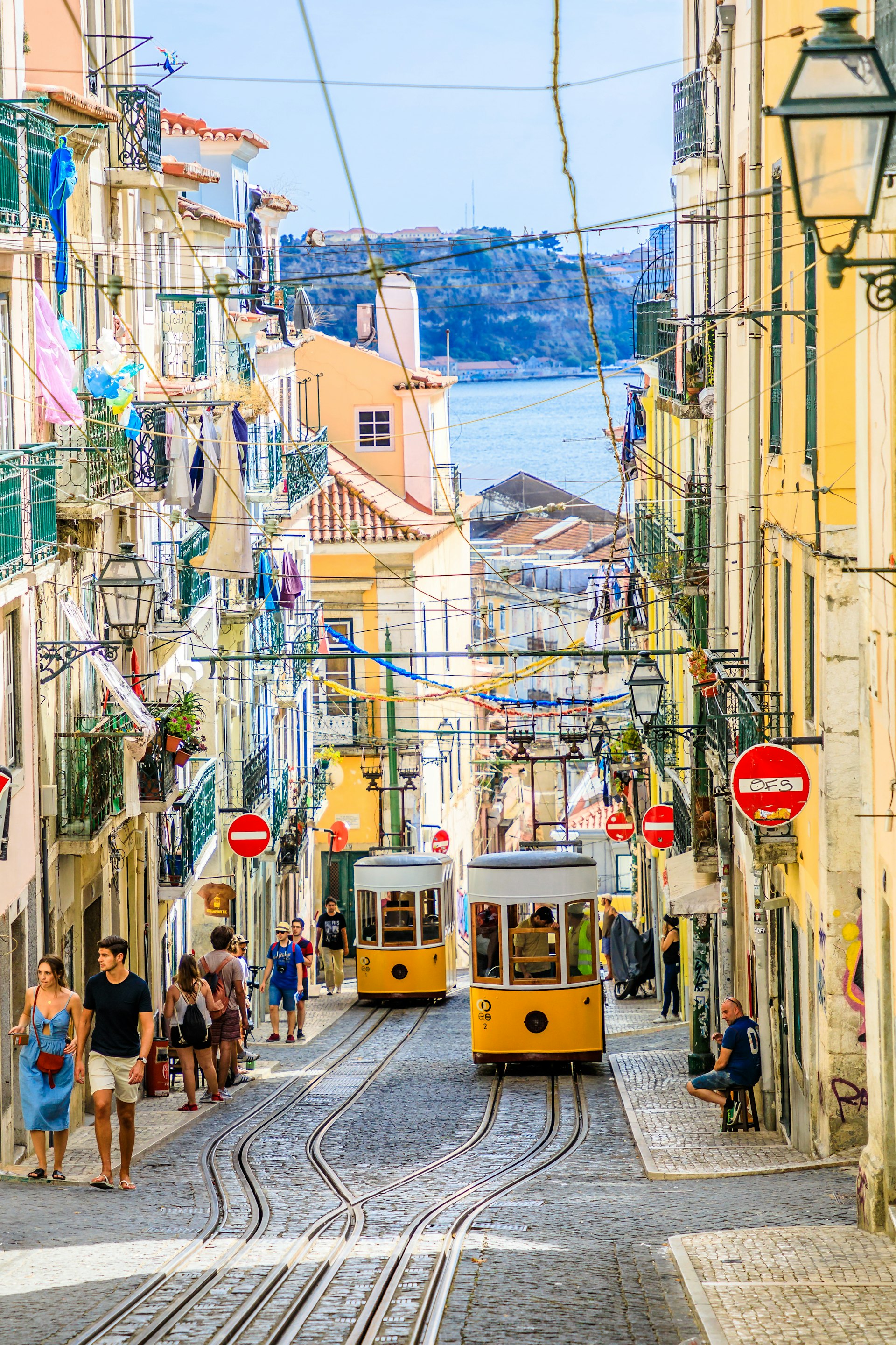 Two yellow trams running down a cobbled hilly street with a body of water in the distance