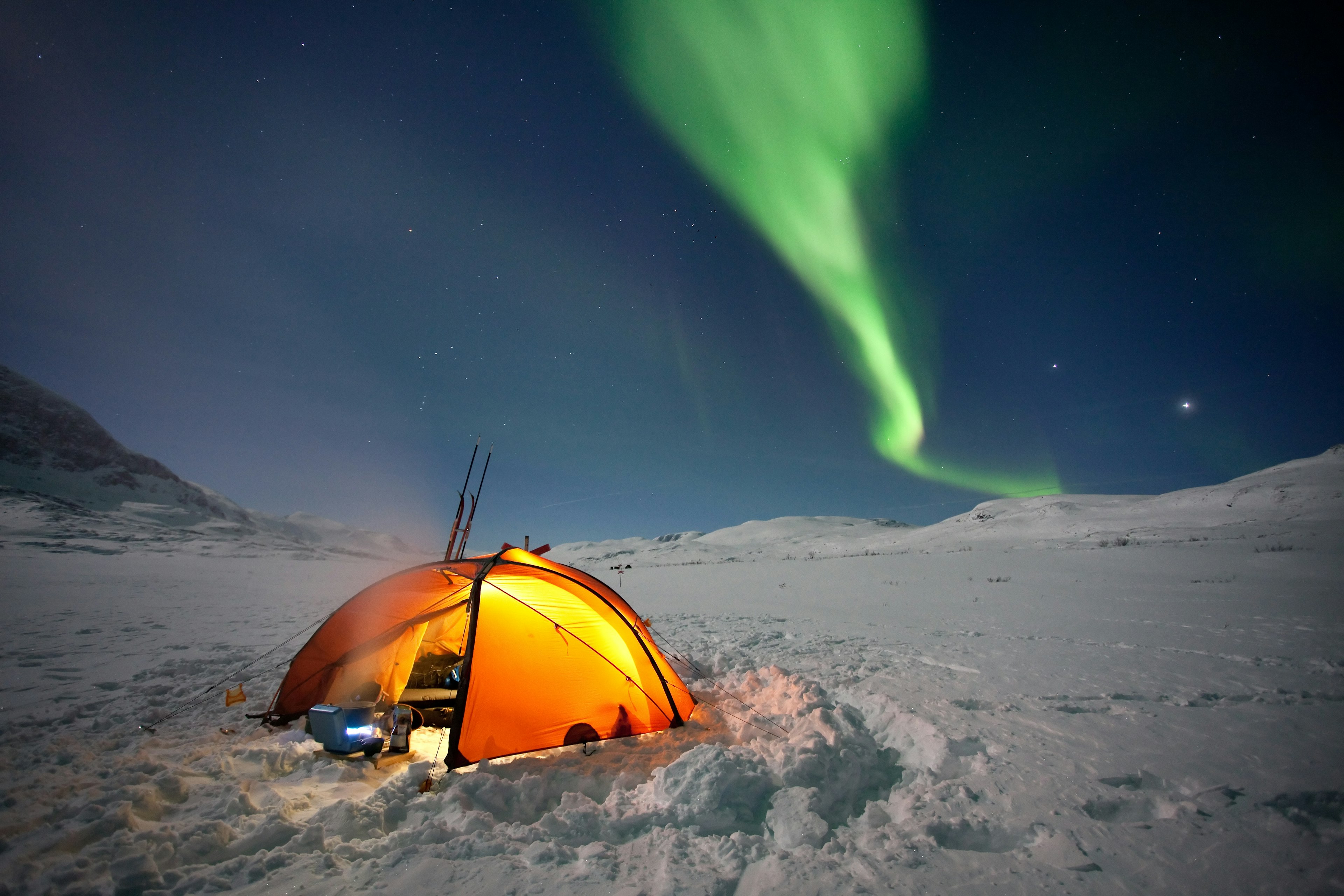 A tent in a snowy field illuminated at night under the Northern Lights.