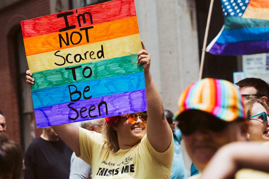 A smiling person at a festival holds up a rainbow-colored sign that says 