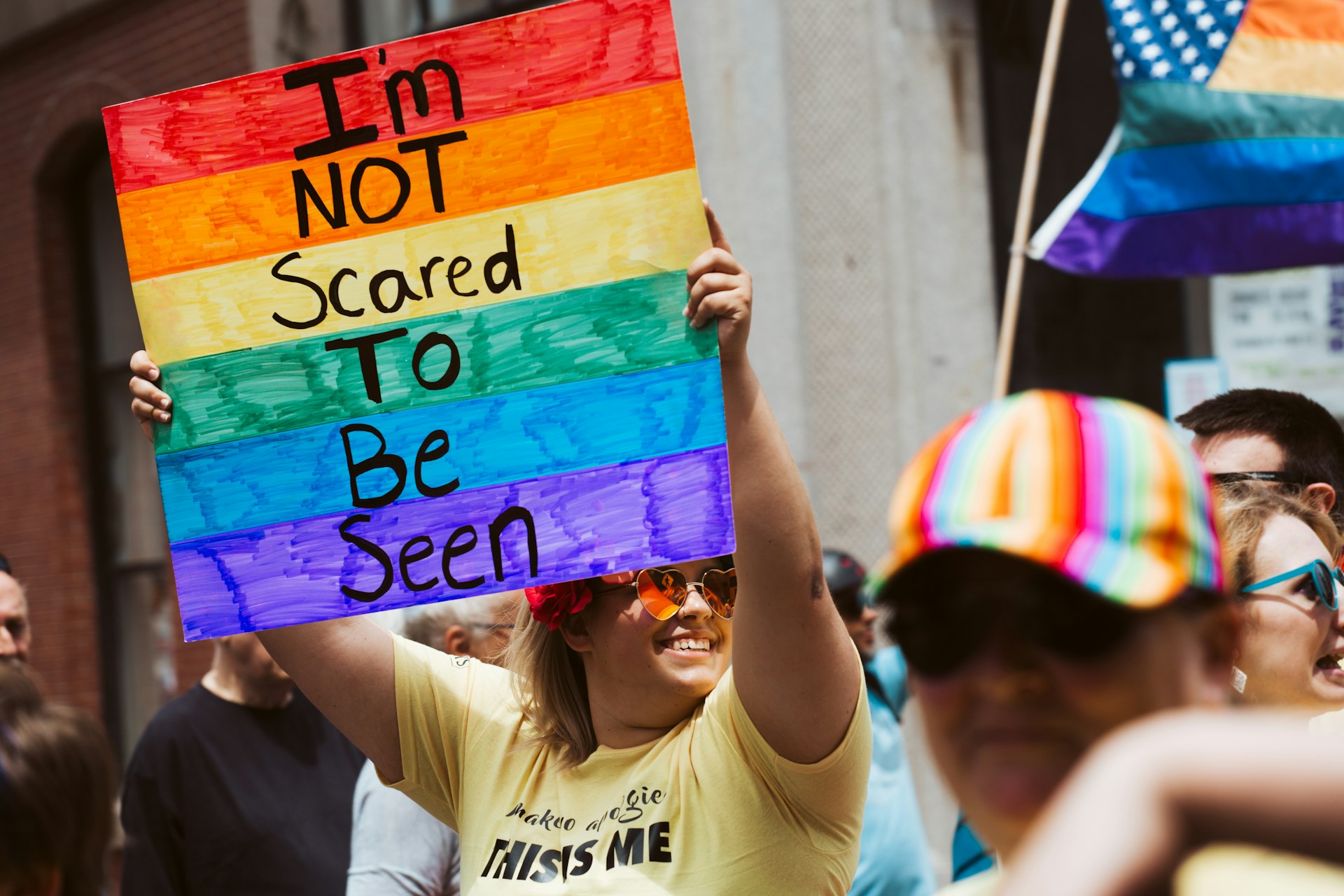 A smiling person at a festival holds up a rainbow-colored sign that says "I'm not scared to be seen"