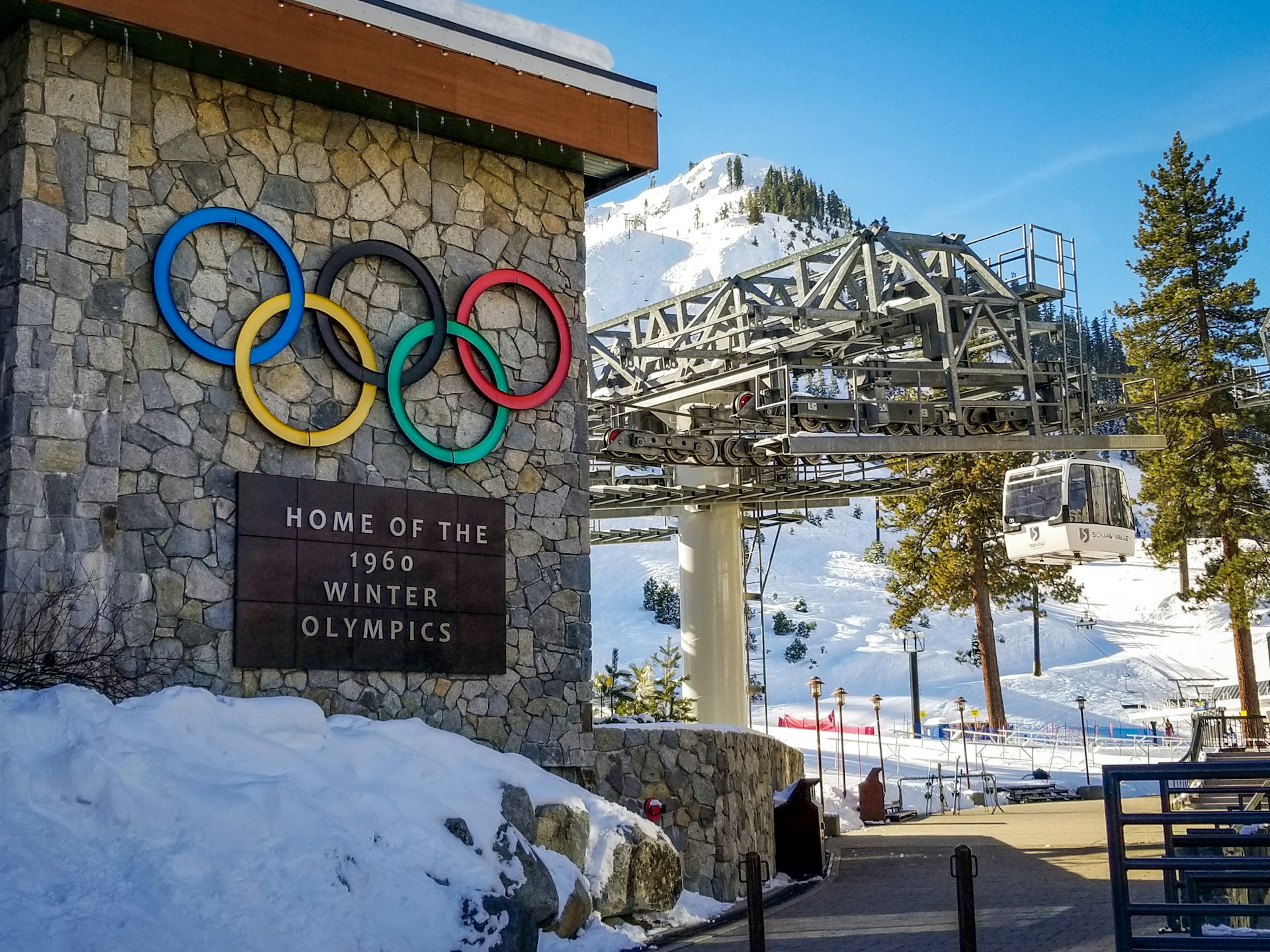 A photo of a resort sign with Olympic rings in the base area, and chairlift in the background