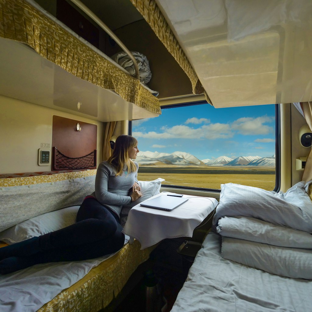 Young woman lies on the bed and looks through the window of the sleeper train crossing picturesque Tibet. Female tourist observing the landscape while traveling along the Trans-Himalayan railway.