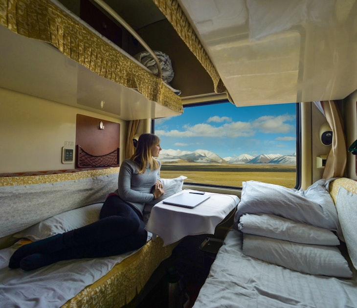 Young woman lies on the bed and looks through the window of the sleeper train crossing picturesque Tibet. Female tourist observing the landscape while traveling along the Trans-Himalayan railway.