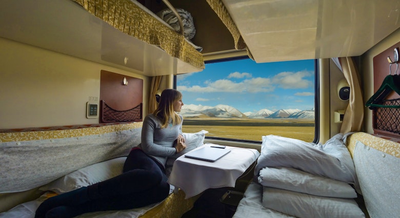 Young woman lies on the bed and looks through the window of the sleeper train crossing picturesque Tibet. Female tourist observing the landscape while traveling along the Trans-Himalayan railway.