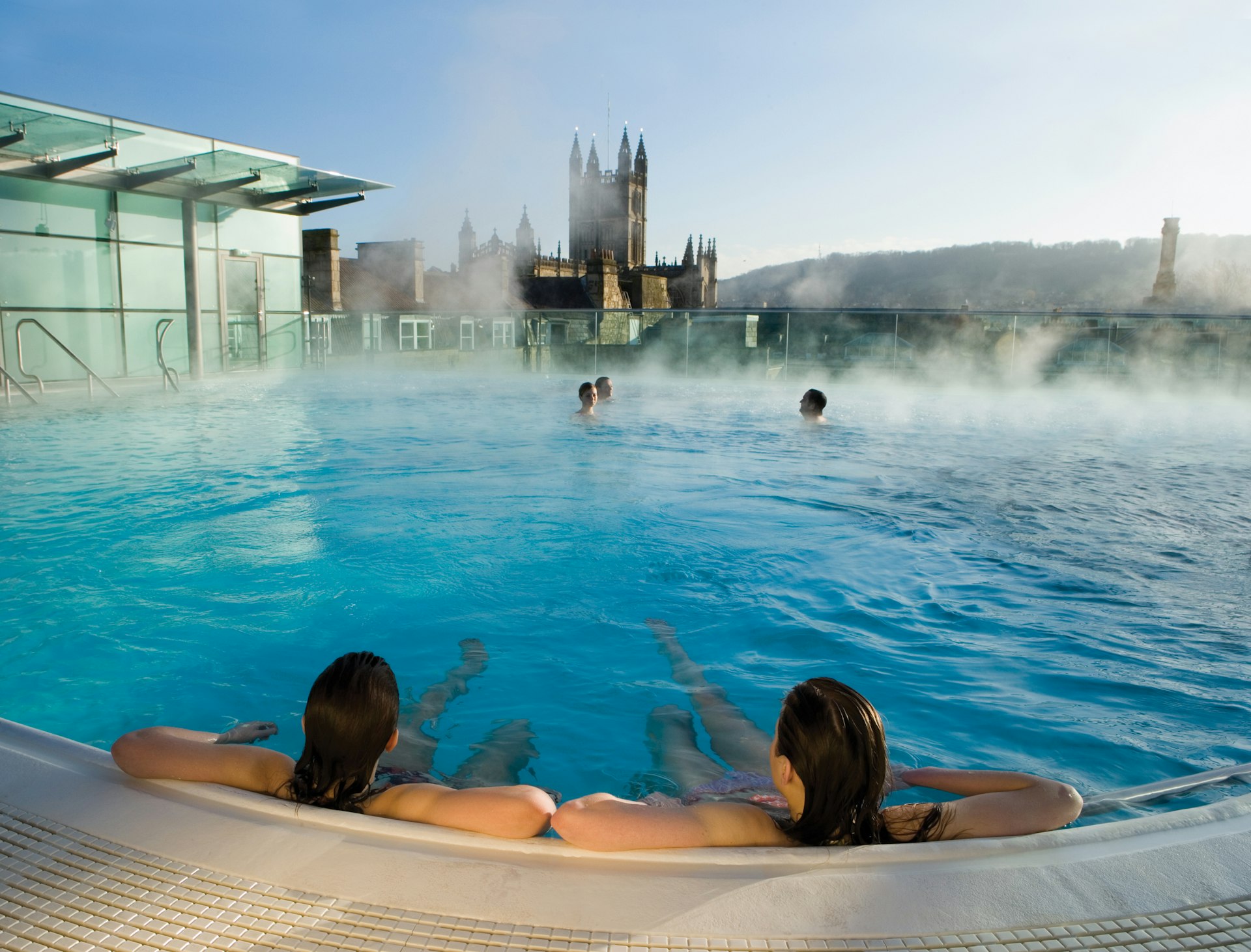 Two people lounge on the edge of a rooftop swimming pool with their backs to the camera. Steam rises from the water. A cathedral building is in silhouette in the distance