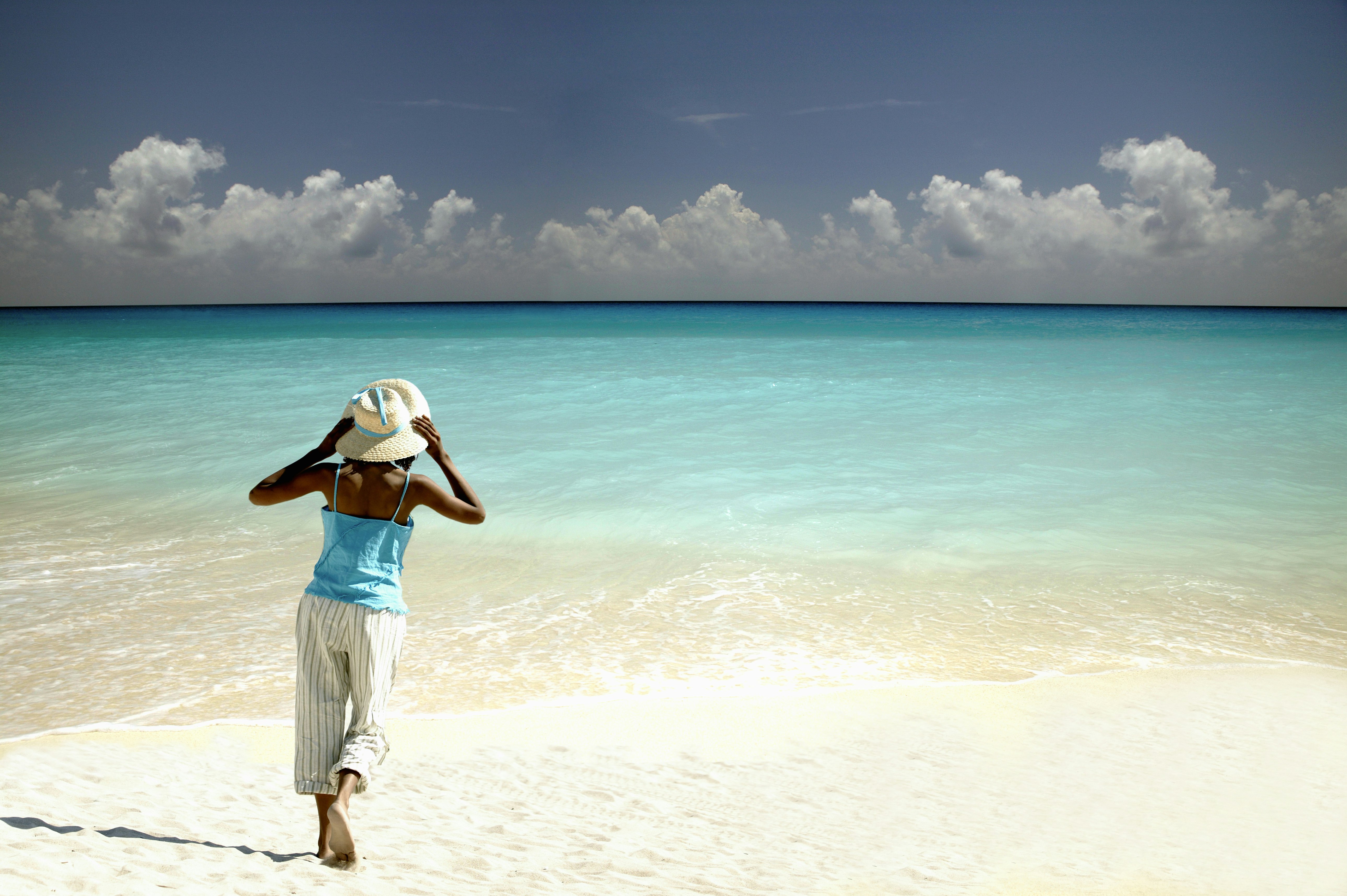 A woman wearing hat on a white-sand beach with turquoise water, seen from behind