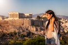 A beautiful Europe traveler woman stands in front of the Acropolis of Athens, Greece, and reads a travel guide book