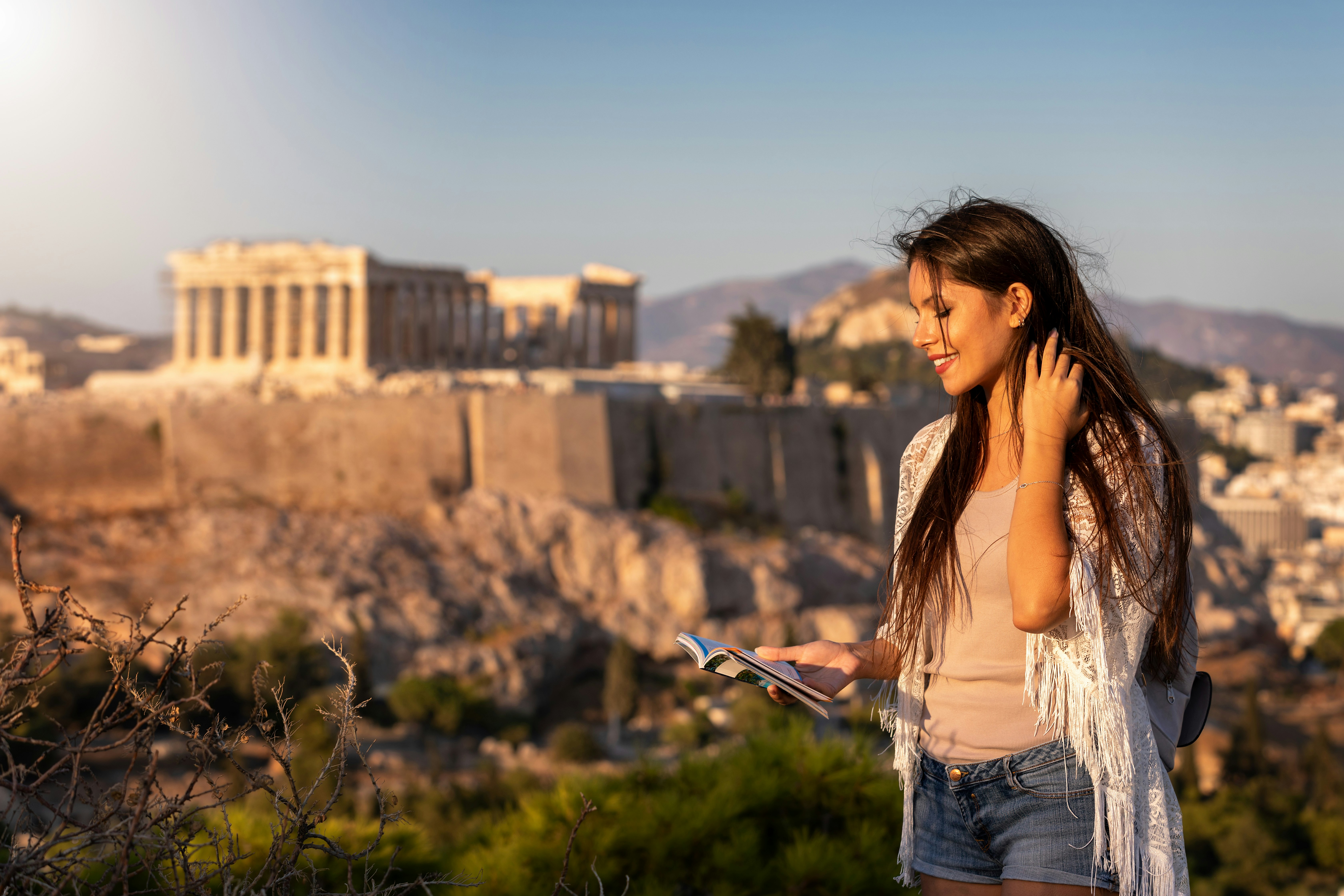 A beautiful Europe traveler woman stands in front of the Acropolis of Athens, Greece, and reads a travel guide book
