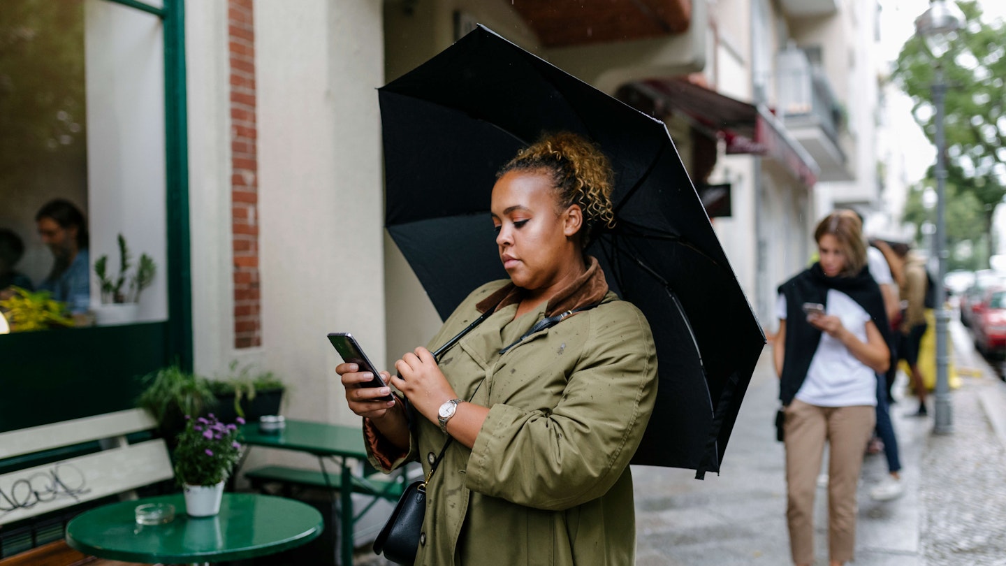 A woman with an umbrella waiting in line, following social distancing guidelines and using her smartphone. 