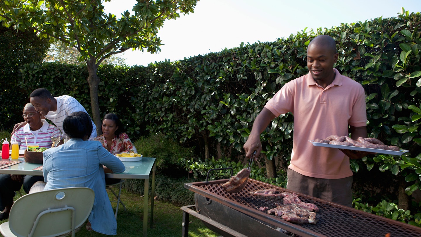 A man is braaing (Afrikaans for barbecuing or roasting) in the garden for a table of guests.