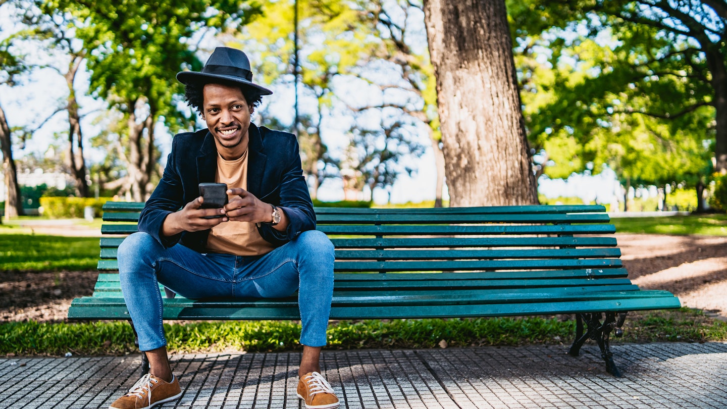 Smiling young African man using smartphone on park bench.