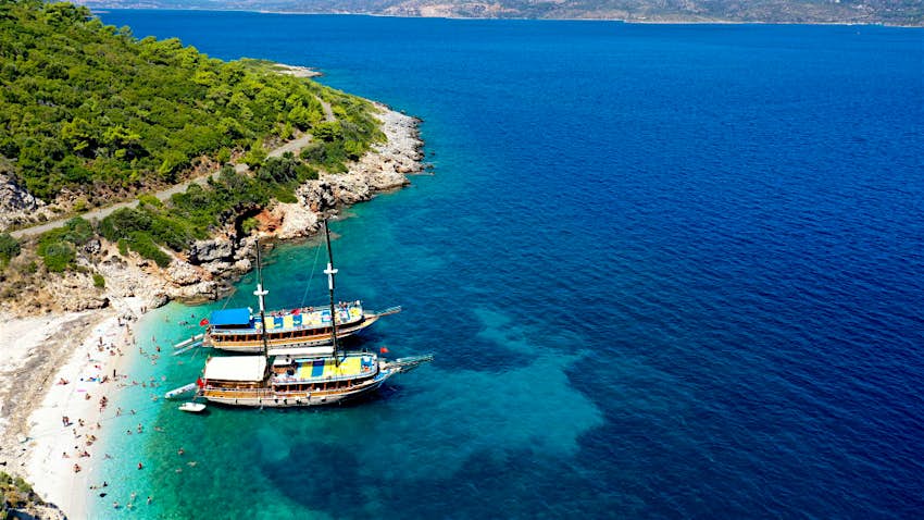 A view from above of one of the small sandy beaches of Turkey's Dilek Peninsula. The small cove is lapped by beautiful blue water, and two large ships are moored nearby. People lounge on the beach and swim in the sea.