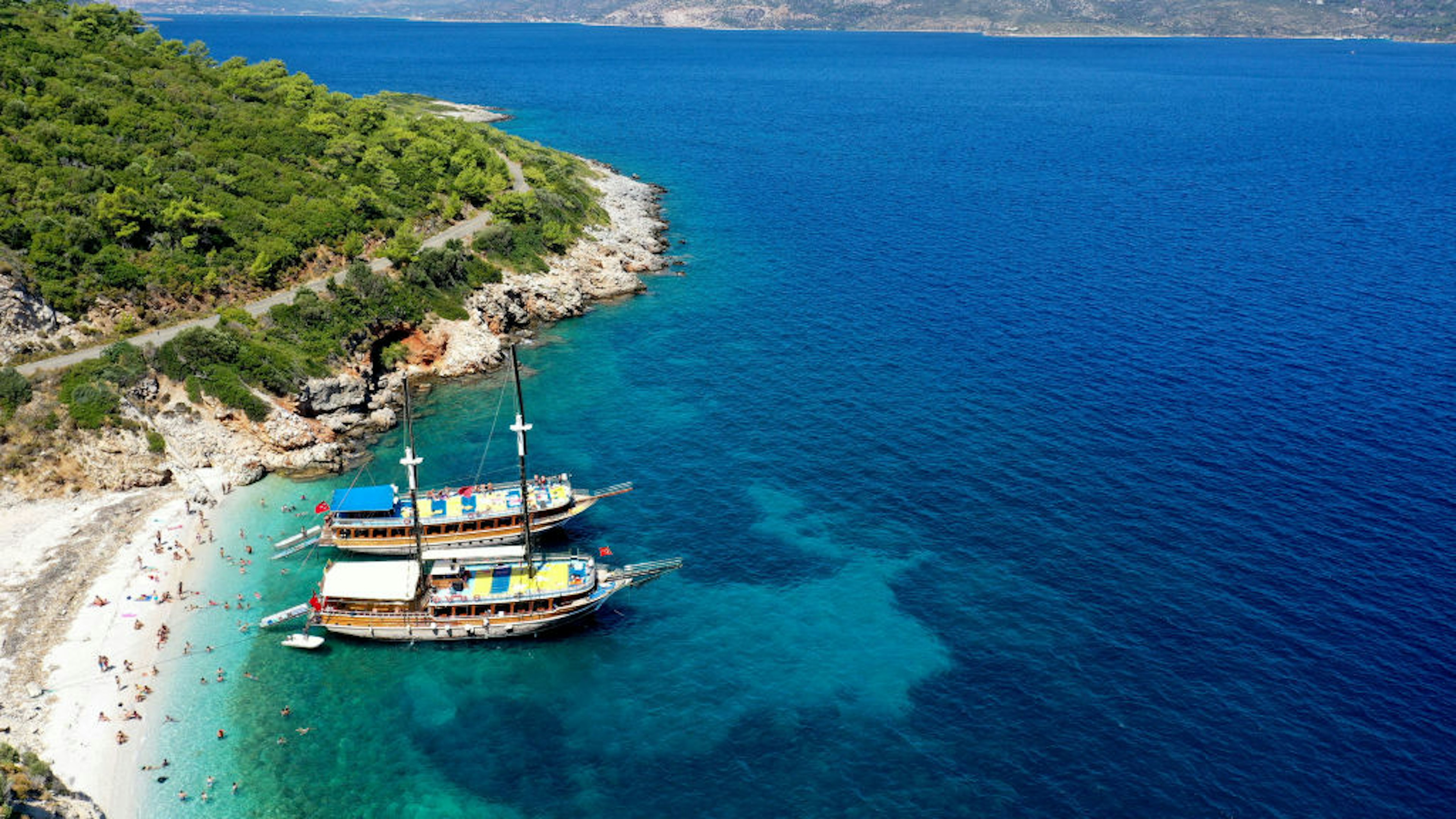 A view from above of one of the small sandy beaches of Turkey's Dilek Peninsula. The small cove is lapped by beautiful blue water, and two large ships are moored nearby. People lounge on the beach and swim in the sea.