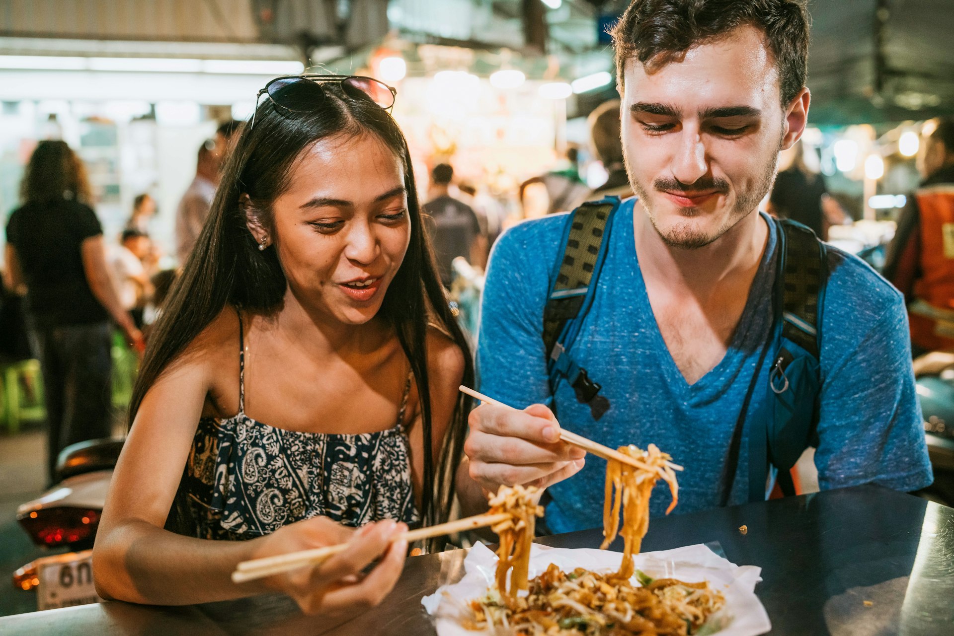 A woman with long hair and a man with a mustache having dinner at a night market 