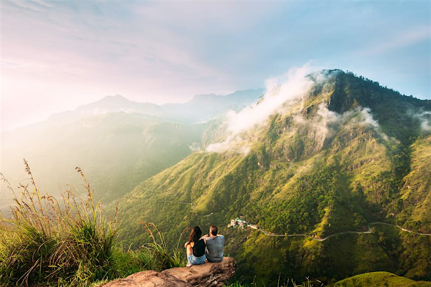 A man and woman sit on a viewpoint in Sri Lanka, looking down over a valley and other mountains. The valley is covered in green forest.
